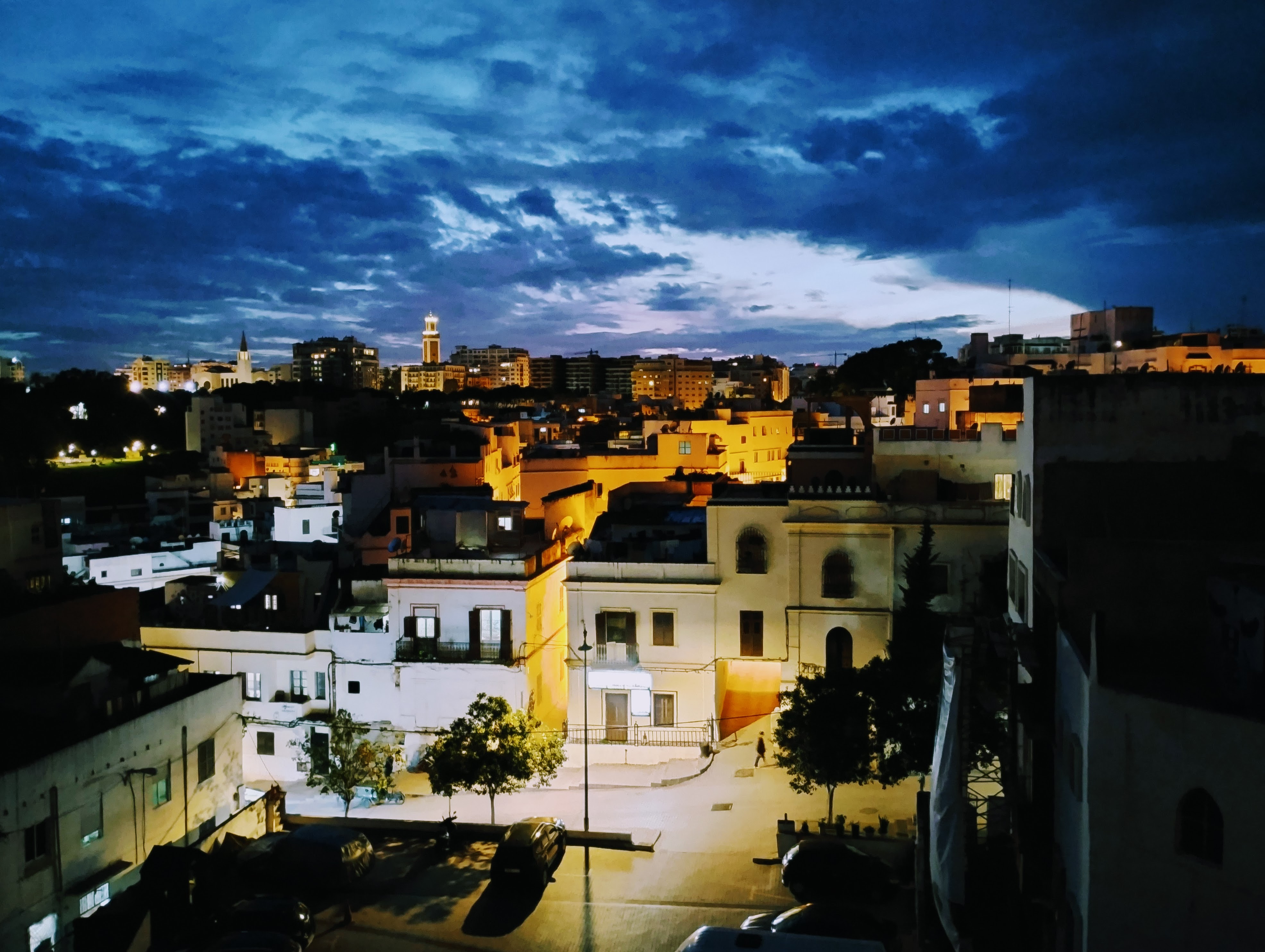A nighttime view of Tangier, with buildings illuminated by streetlights and interior lighting, under a dramatic, cloudy blue sky.