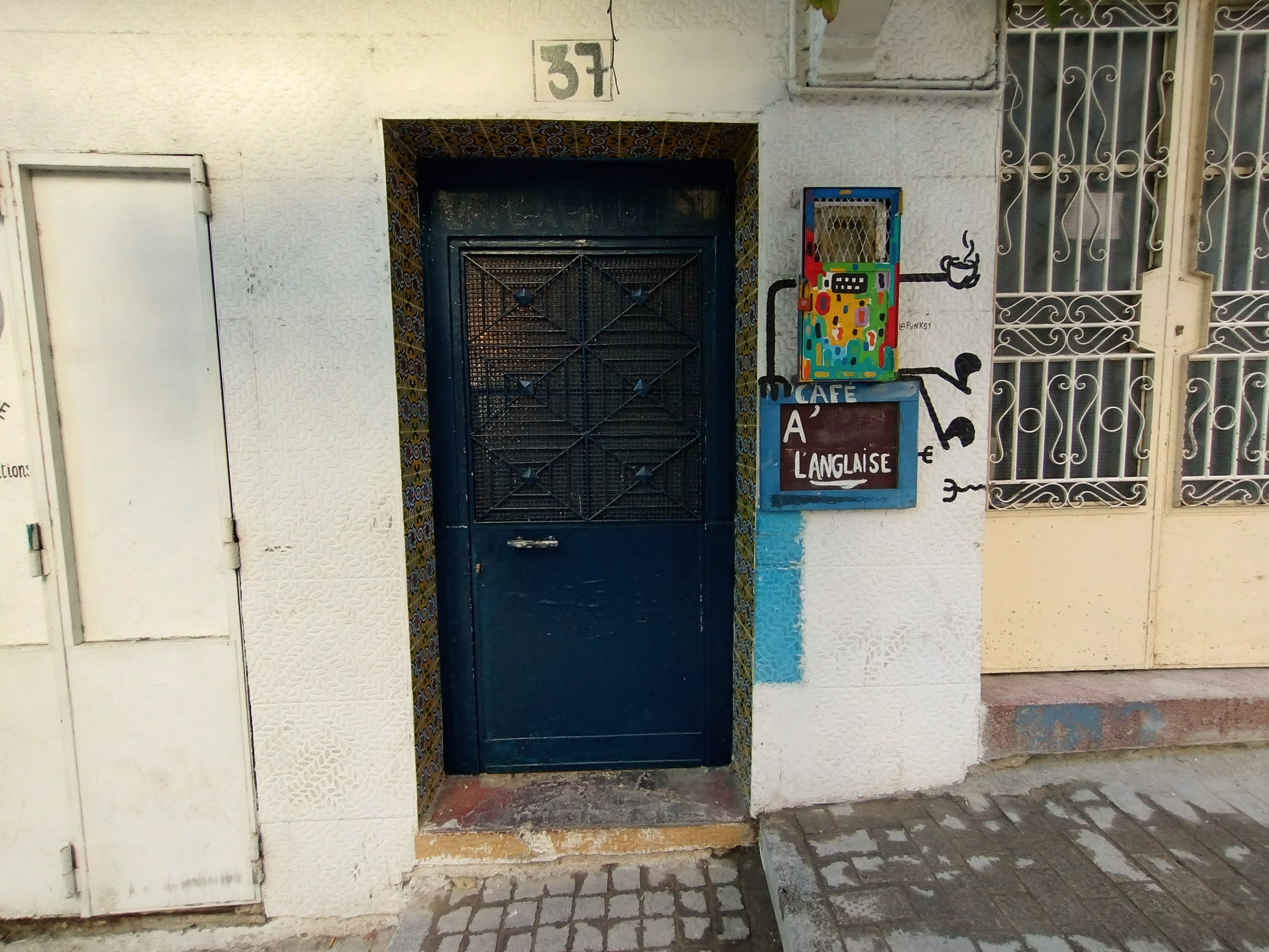 Street view of a building façade with a dark blue metal door, a painted sign for "Cafe A' L'Anglaise", and an electricity meter disguised to look like it is holding a cup of coffee.