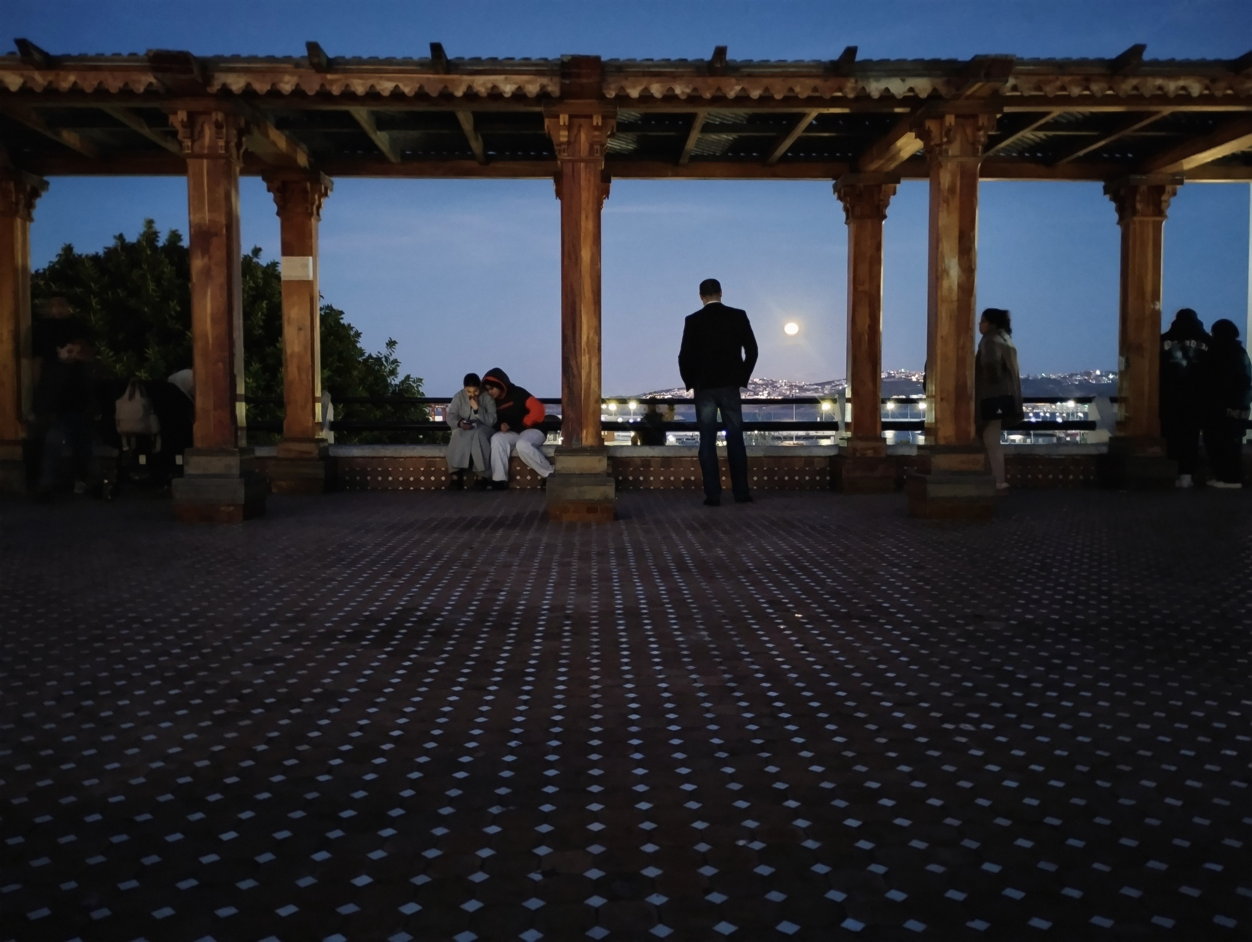 A full moon rises at night through a wooden pergola. A man in a black jacket stands with his back towards the camera, between two beams. Several people are gathered in the background, some sitting, some standing, with a patterned floor visible in the foreground.