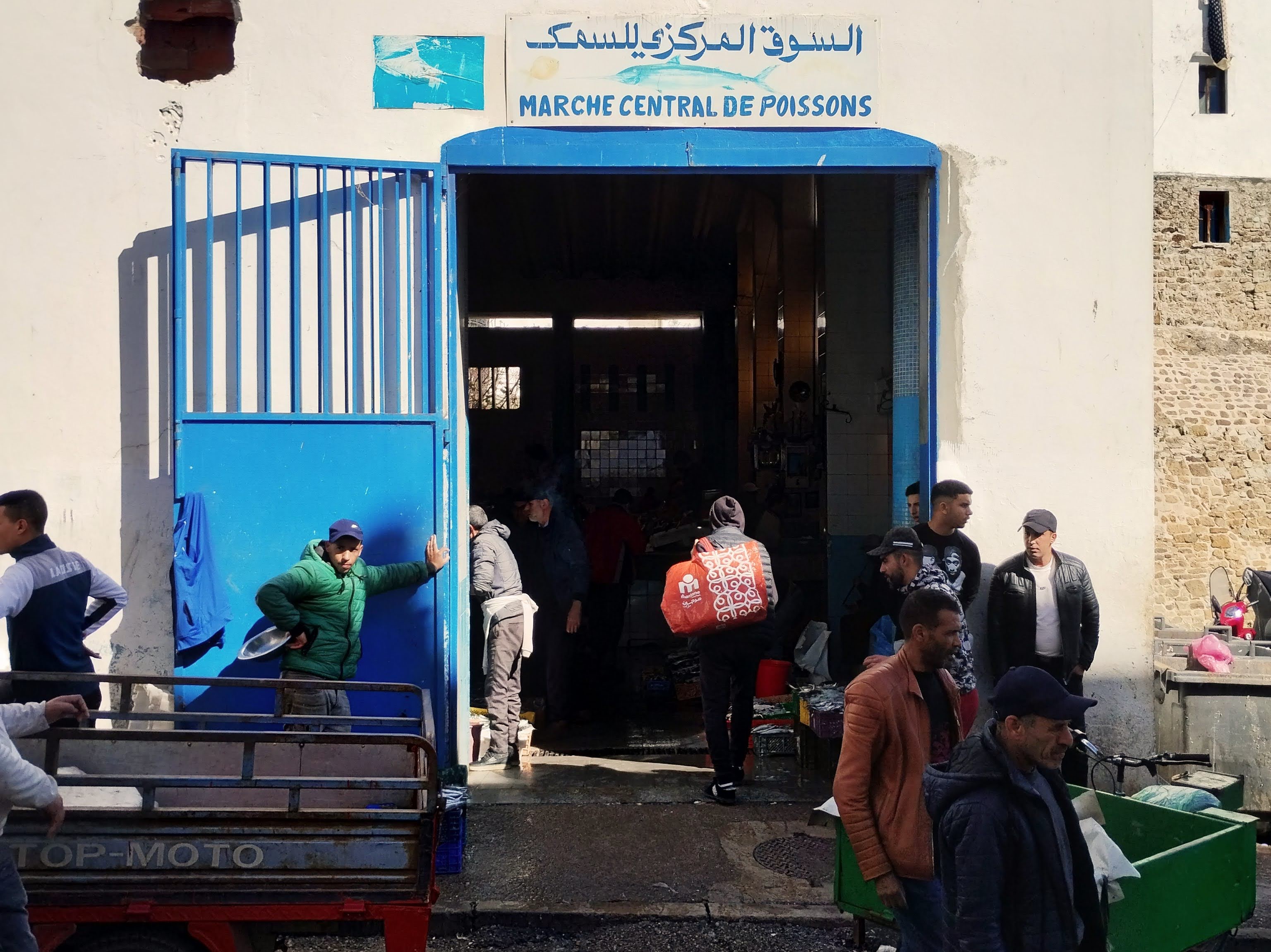 Exterior view of a bustling fish market entrance, marked by a blue archway with signage in Arabic and French; people gather in the doorway and adjacent to the entrance, including men wearing jackets and caps, while others move in and out, some with baskets, and a delivery truck is parked in front.