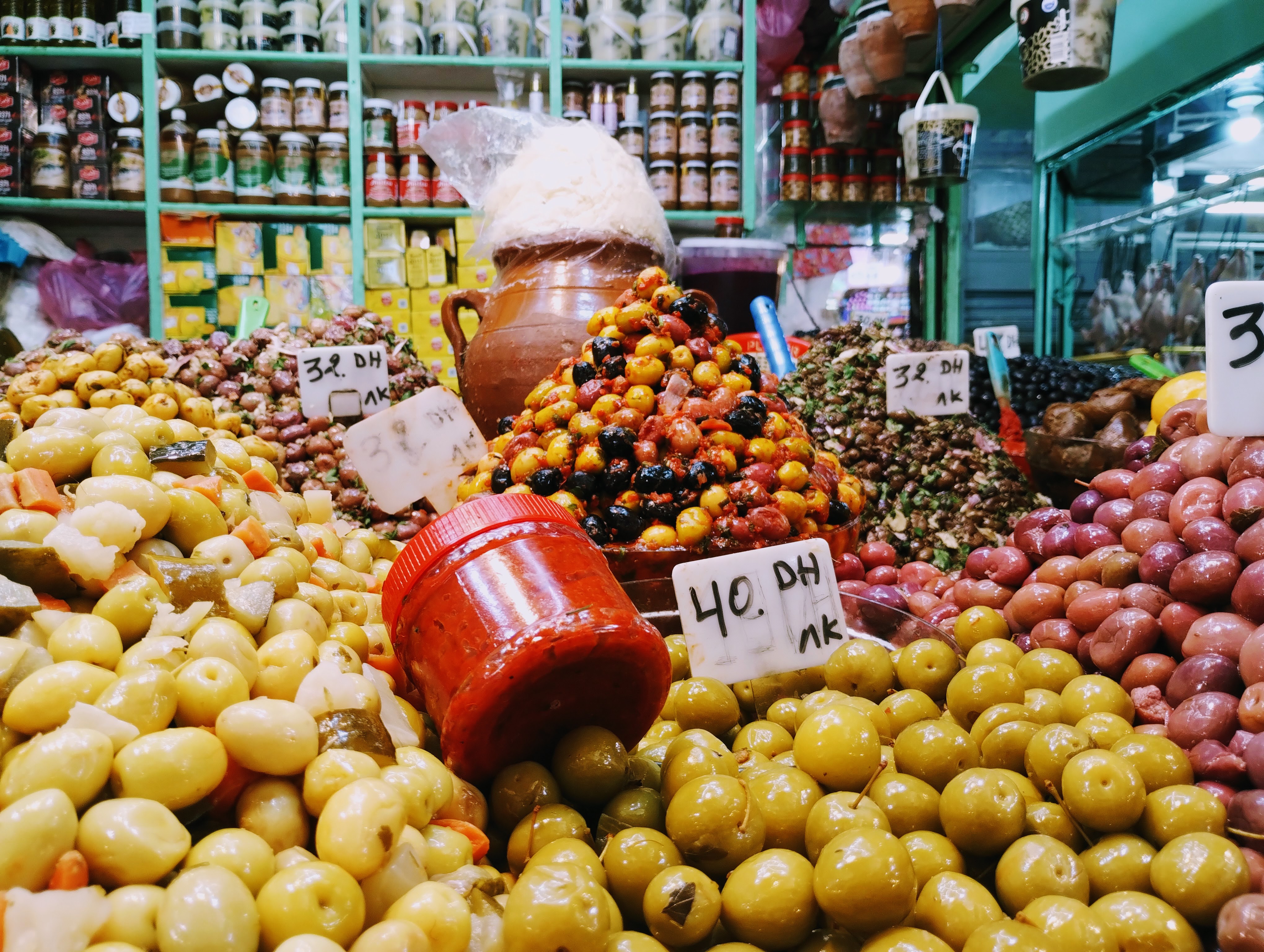 On a vibrant market stall, a jar of red harissa sits amid a colourful array of green, red, and black olives pickled vegetables with handwritten price tags