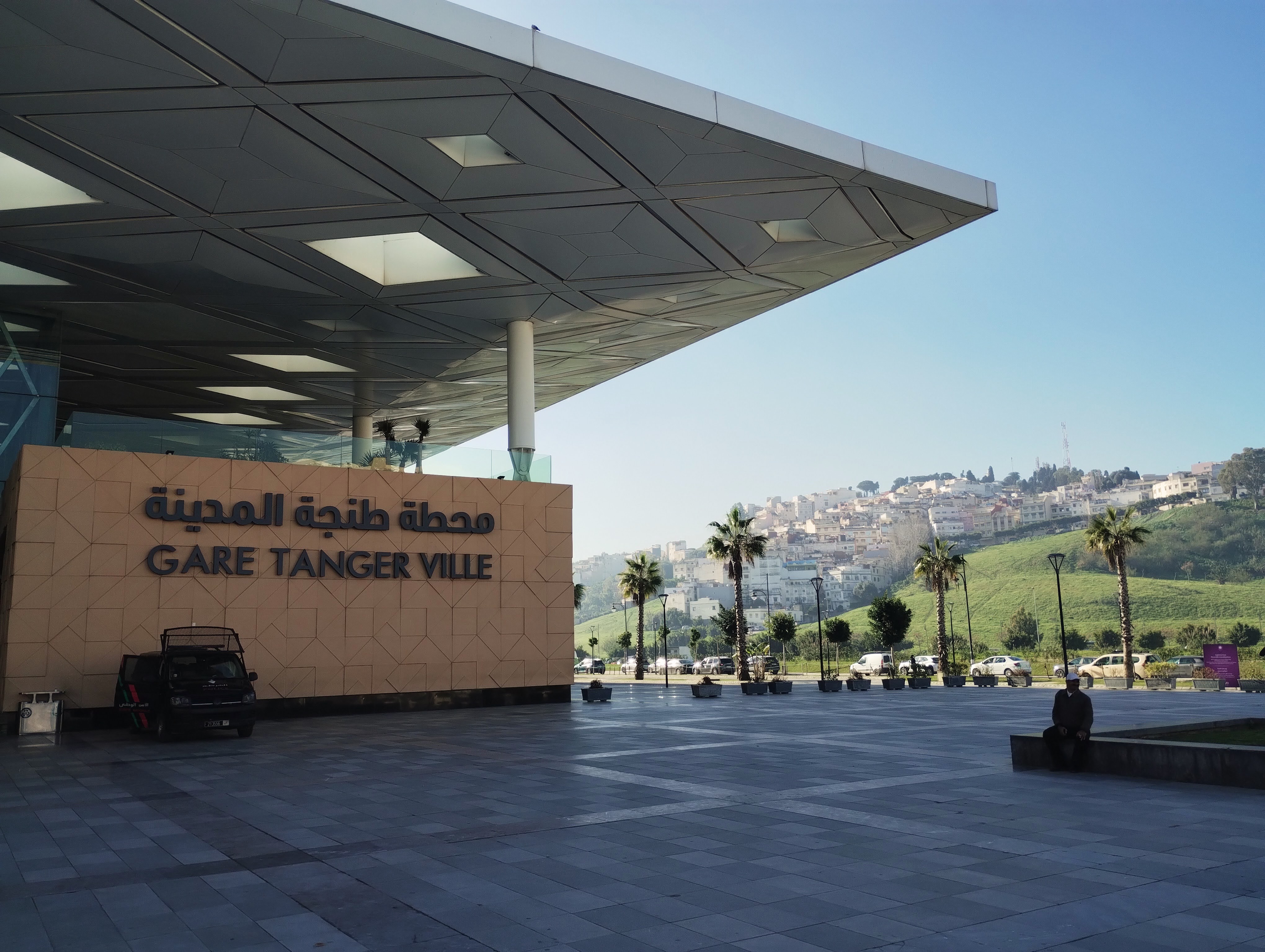 Exterior of Gare Tanger Ville, a modern train station in Tangier, Morocco, with a tan facade and geometric roof overhang, with text in Arabic and English. In the background is a city-scape on a hillside, and palm trees.