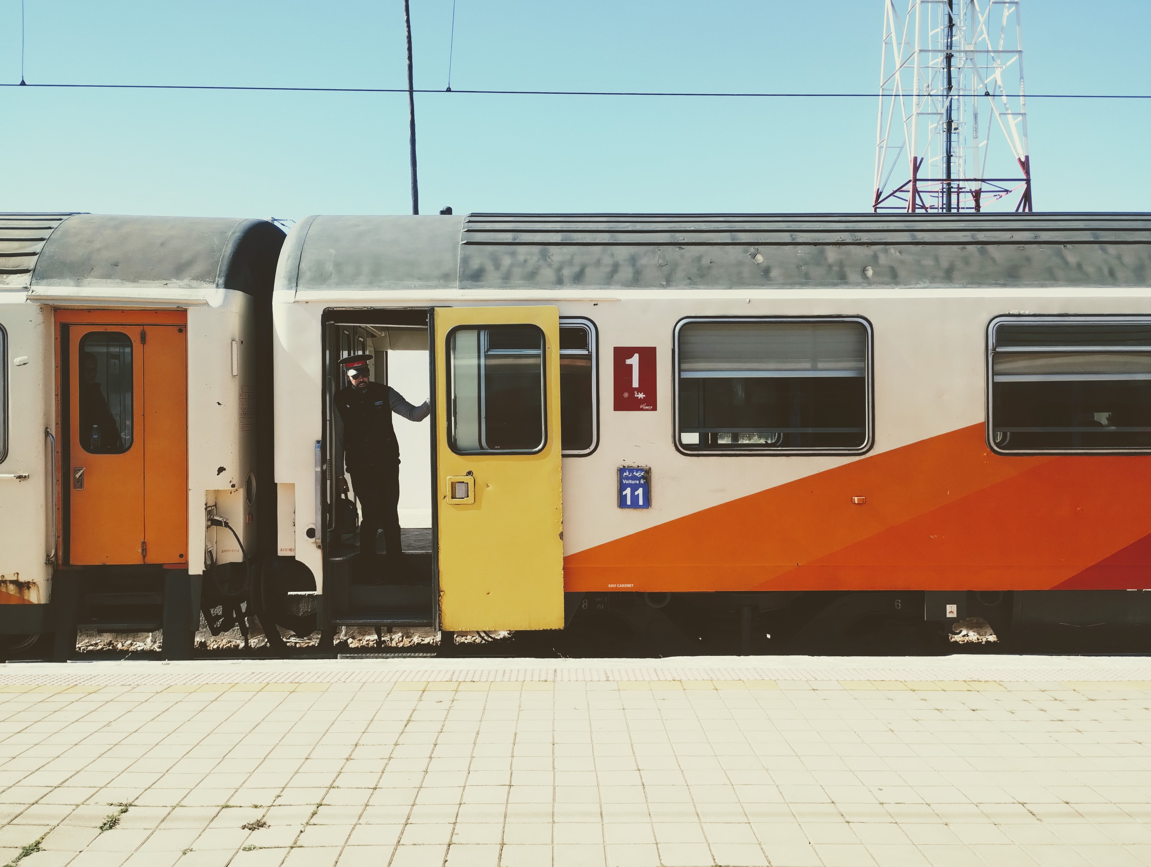 Side view against a clear blue sky of an ONCF passenger train with a yellow open door, in which the guard is standing. The car's exterior is cream with orange and red accents. Windows are visible and the red number "1" for first class is above the coach number, 11, indicated in white on a blue panel.