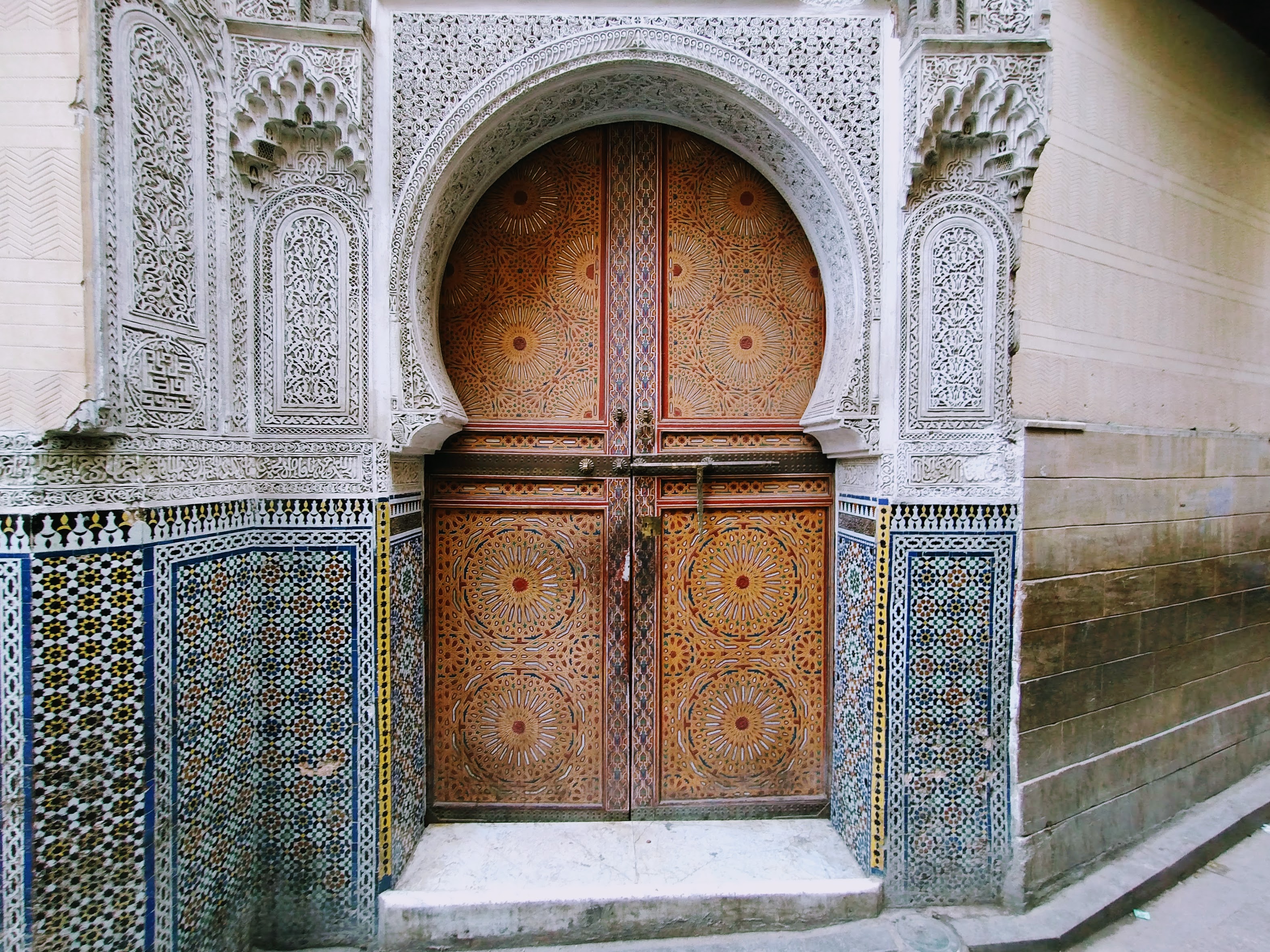 Ornate wooden doors with intricate circular designs set within a decorative horseshoe-arched doorway, flanked by detailed white plaster-work and zellije tiled borders.