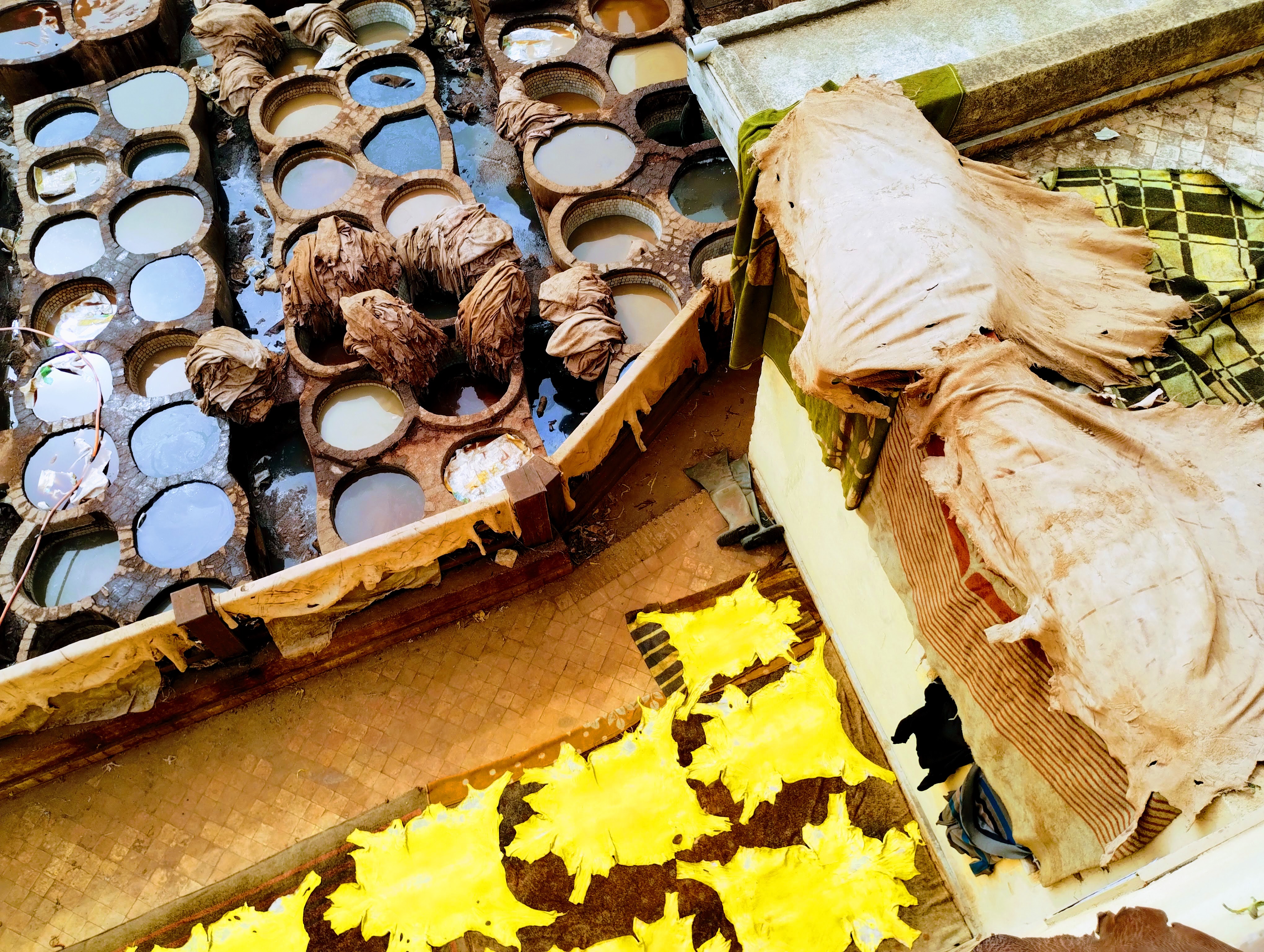 Overhead view of a traditional tannery with numerous circular vats filled with dyes, surrounded by large animal hides drying in the sun. The hides are draped over structures or spread out, and some are a vibrant yellow.