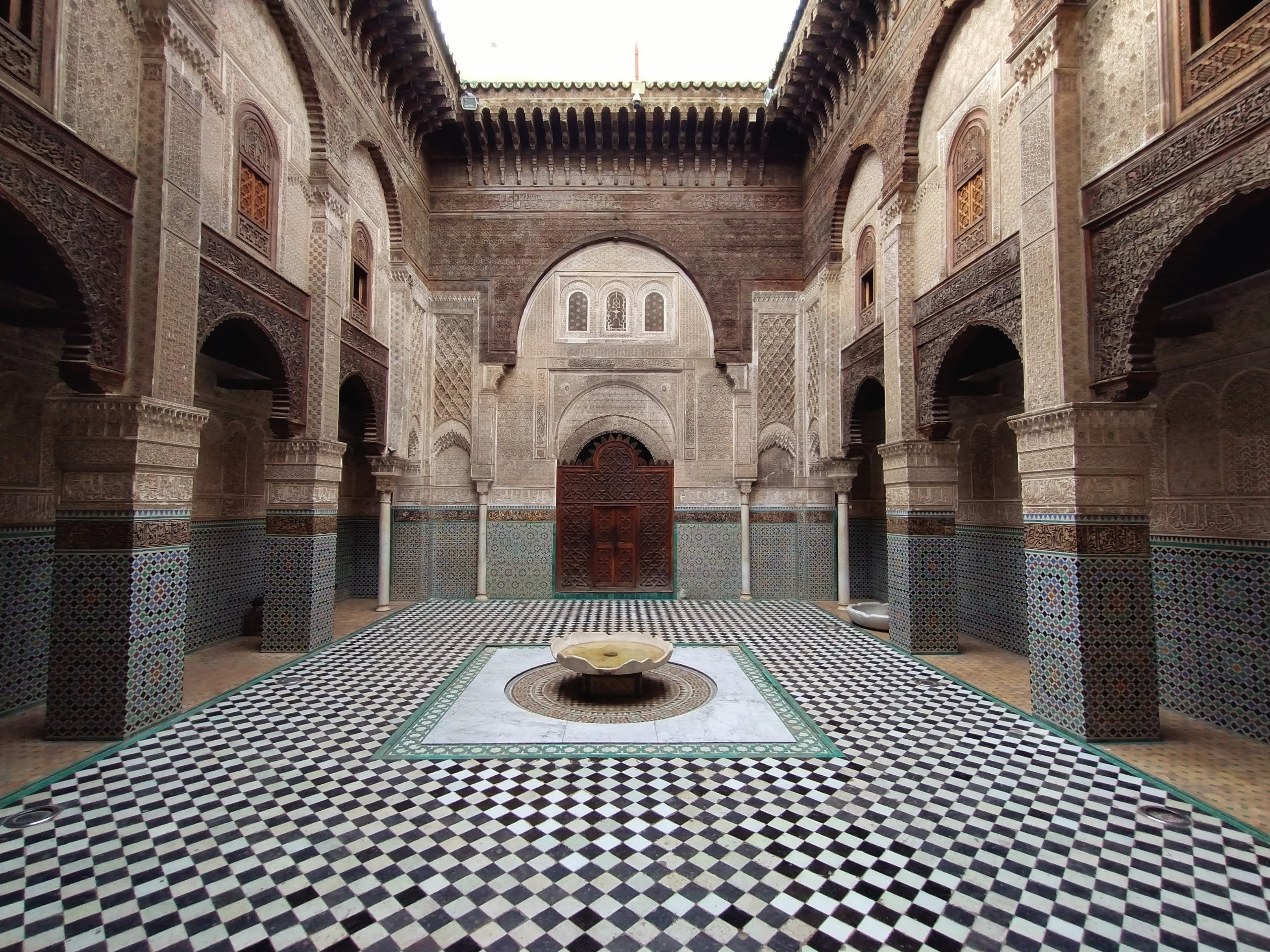 In a courtyard, a fountain surrounded by tiled flooring and pillars decorated with colourful tiles and ornate wooden carvings.