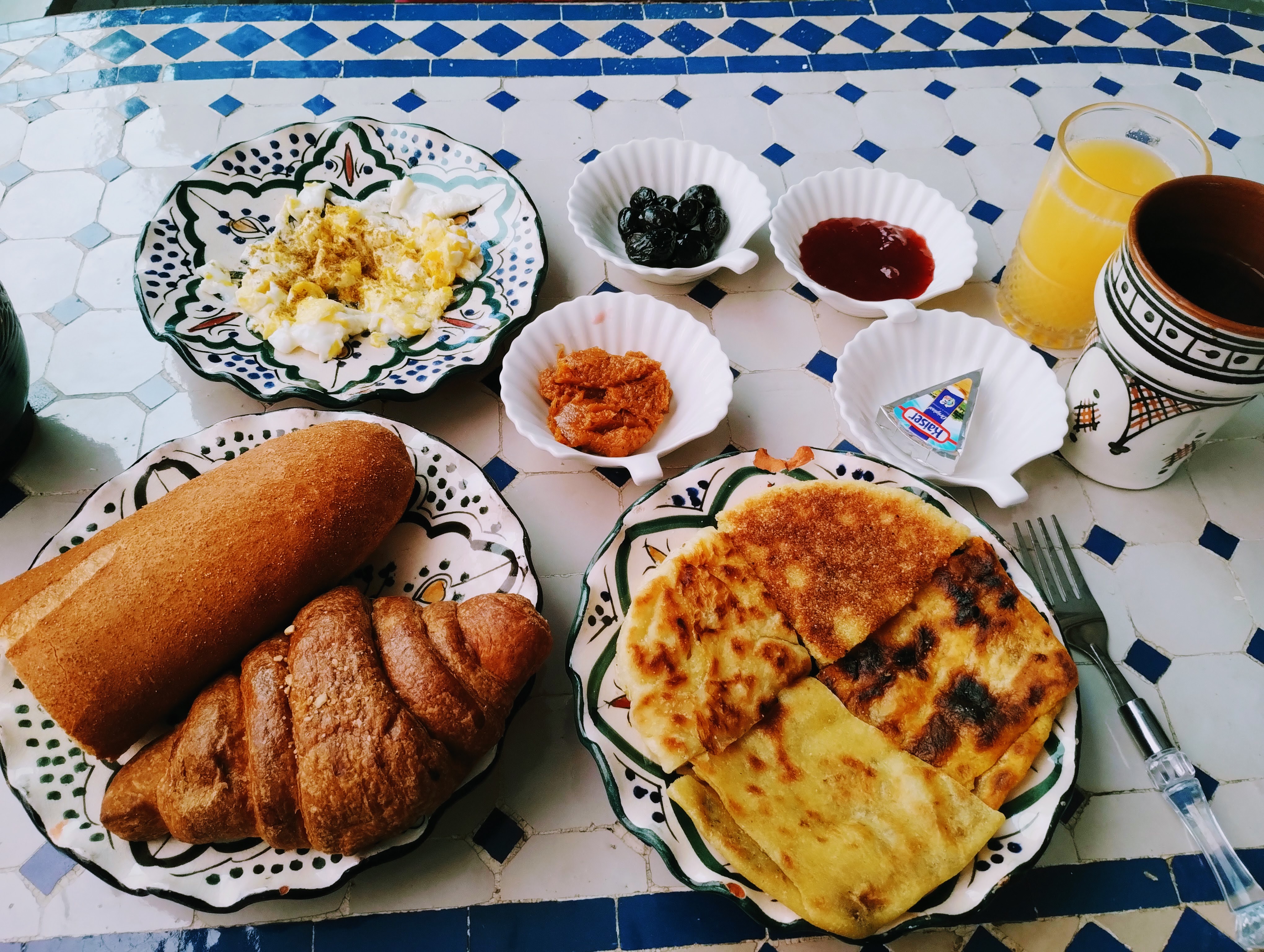 A breakfast of eggs, flat breads, olives, baguette and croissant served with orange juice and coffee on a blue and white mosaic table.
