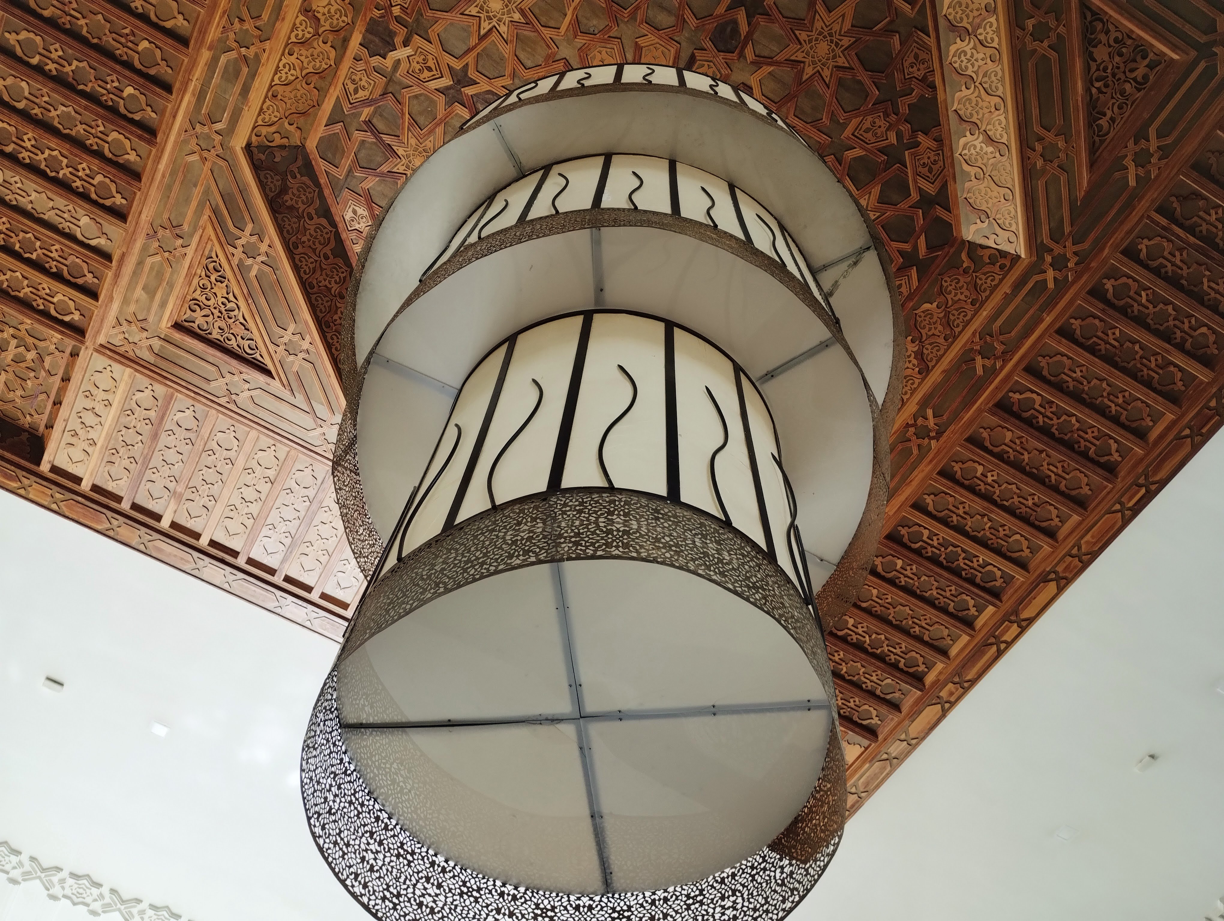 Low-angle shot of an ornate three-tiered chandelier hanging from a carved wooden ceiling with intricate geometric designs and patterns.