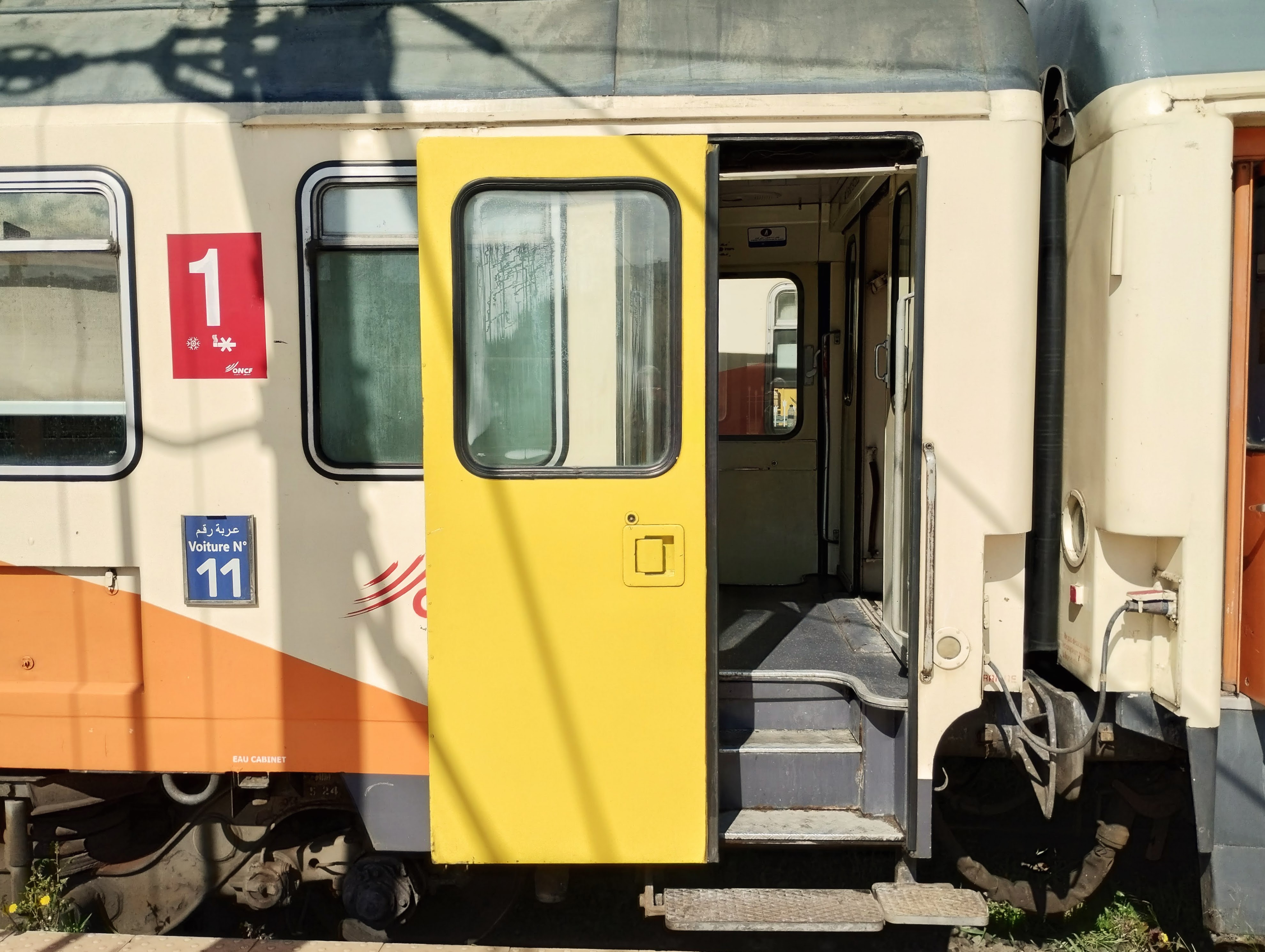 Exterior of a train with an open yellow door revealing the vestibule. The side of the car is cream and orange with a window and number signage.