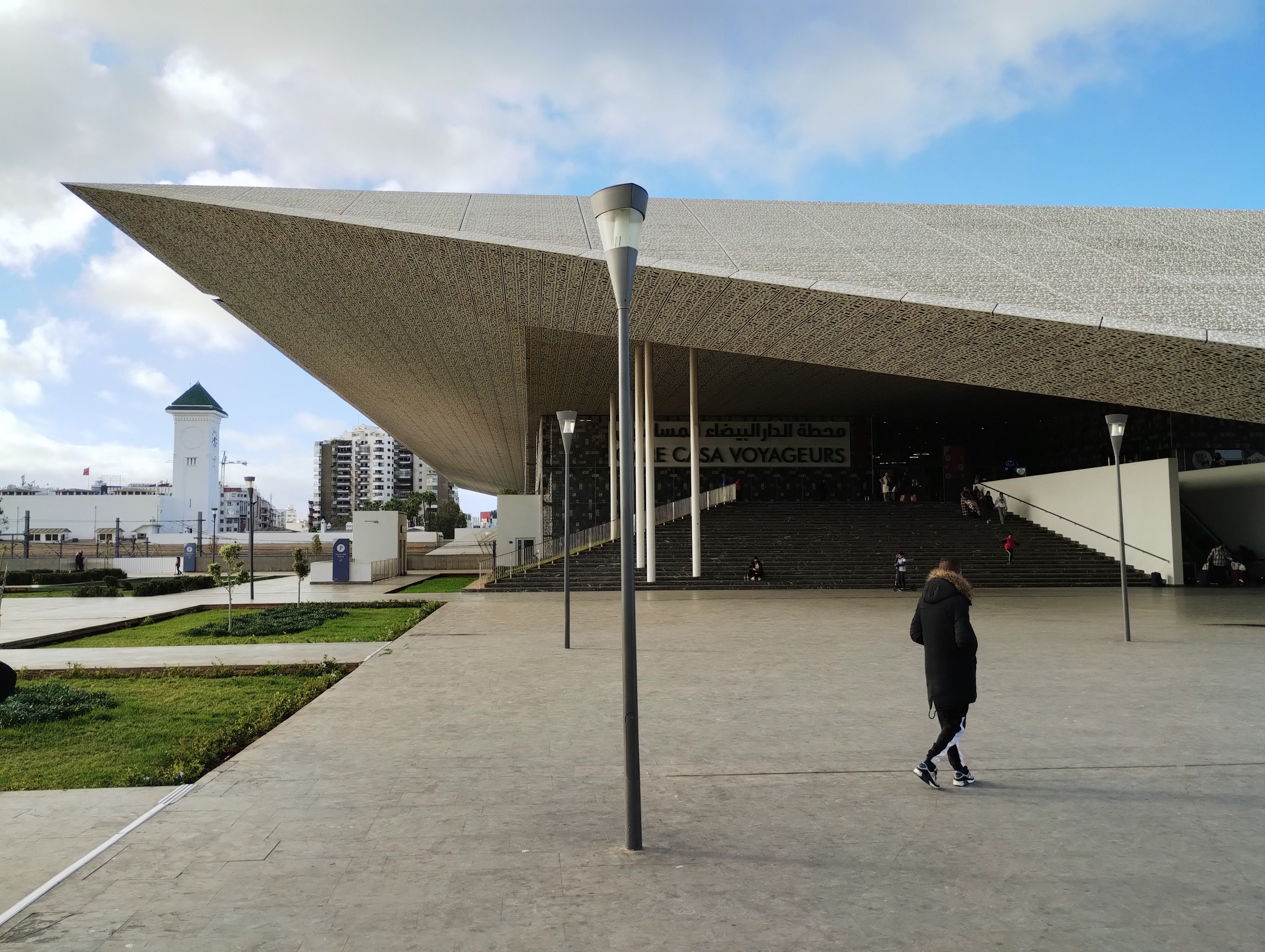 Modern architecture of Casablanca Voyageurs train station, featuring an expansive, geometrically angled roof with patterned texture, a large set of stairs leading to an entrance, and a person in the foreground wearing a long parka with furry hood.