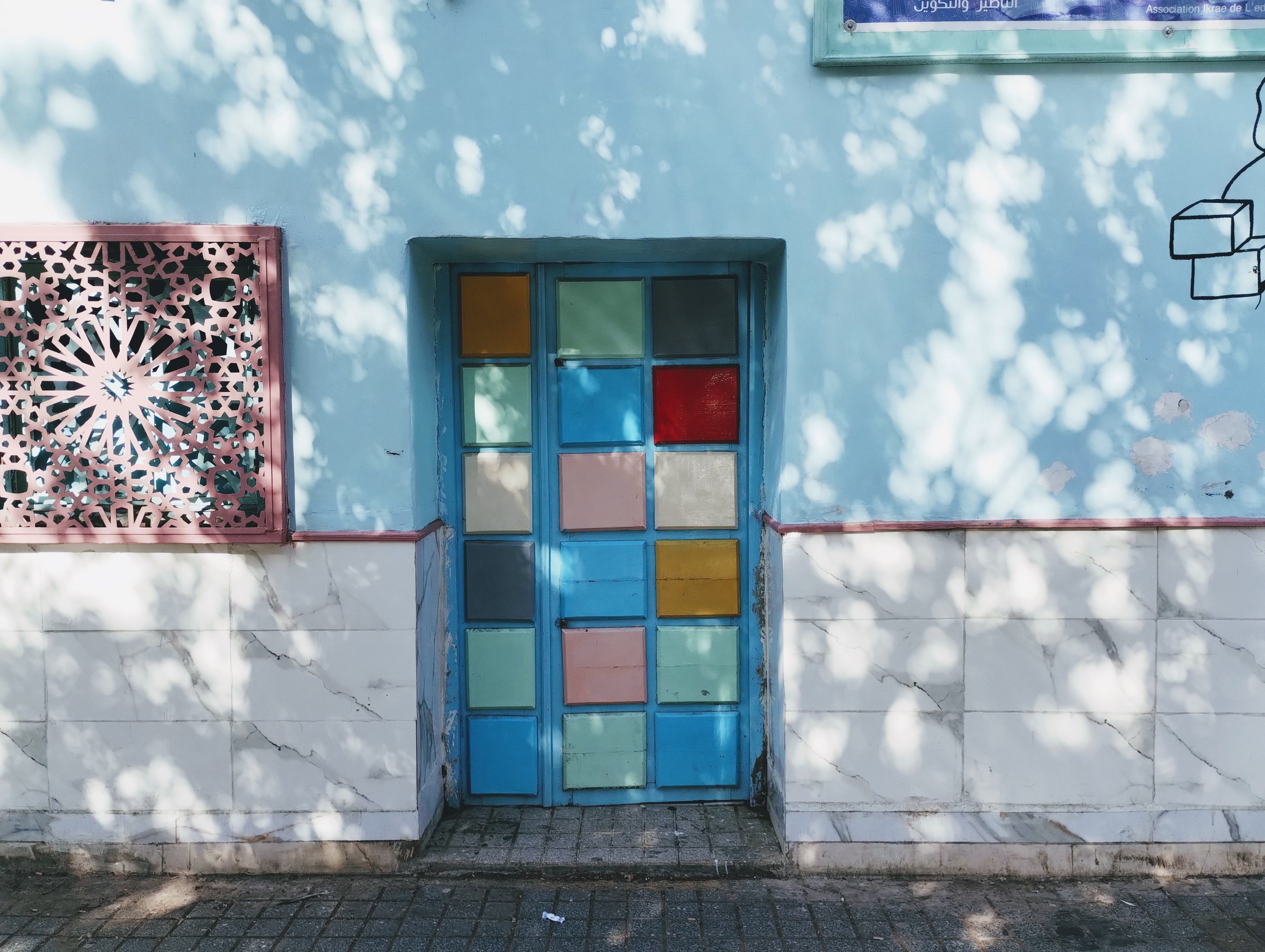 A light blue wall with a blue door and window featuring a colourful mosaic of square glass panes. To the left is a pink latticework window and the lower section of the wall is tiled with white marble-like tiles. The wall is also decorated with the shadow of trees. Above the door is a light blue plaque.