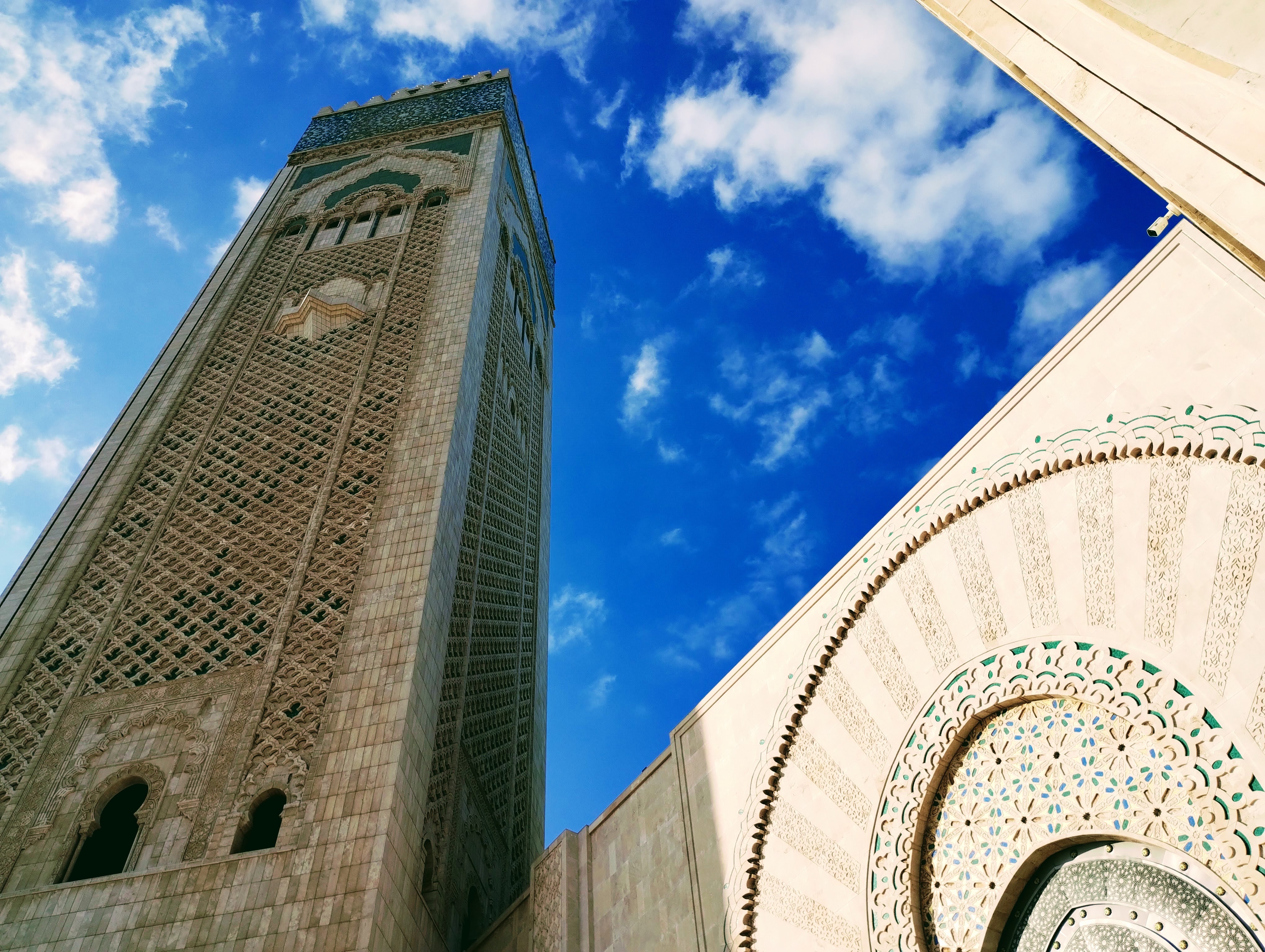 Low-angle shot of Hassan II Mosque in Casablanca, Morocco, featuring a towering minaret with intricate stone carvings and the decorative arched facade of the building against a bright blue sky with scattered clouds.
