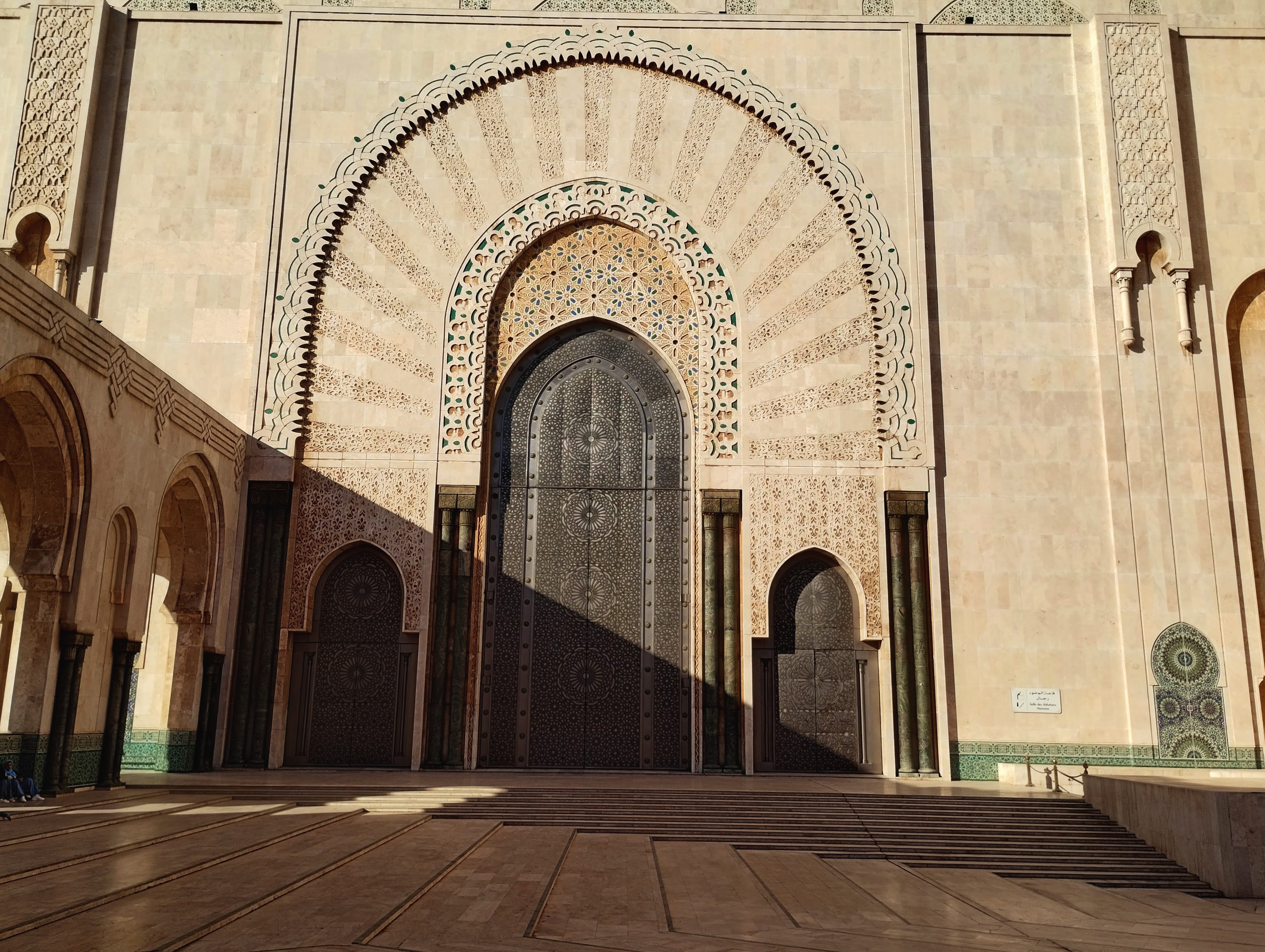 Exterior shot of the Hassan II Mosque in Casablanca, Morocco, featuring the intricately decorated central entrance with a large arched doorway above two ornate, dark metal doors, set within a cream-colored stone facade and flanked by matching columns and smaller archways, with sunlight casting sharp shadows across the mosque's courtyard floor.