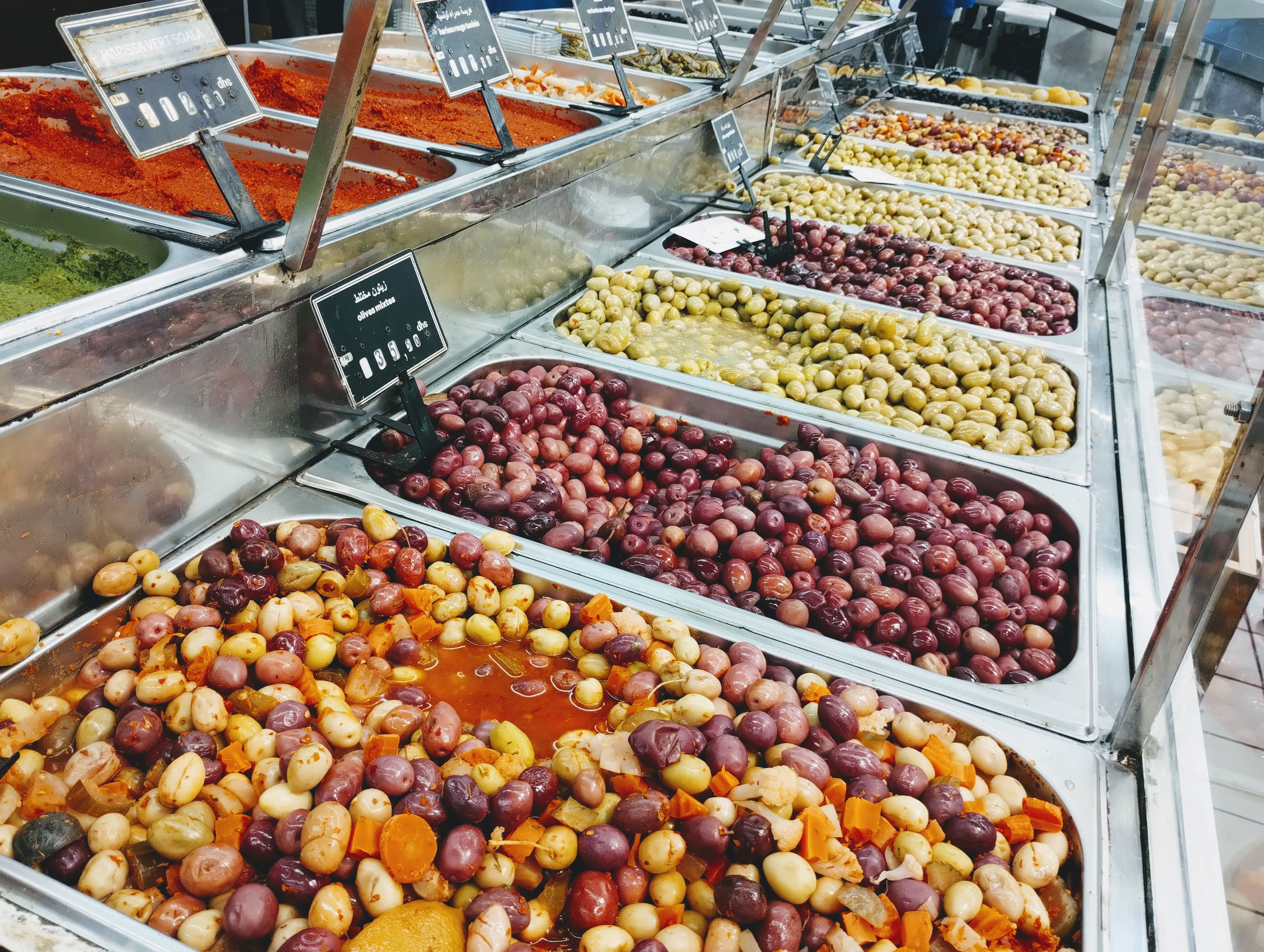 A brightly-lit display of olives and spices in a supermarket. Stainless steel containers hold various olive types, including "olives mixtes," "harissa vert scola," and others labelled with prices and Arabic text. The olives range in colour from green to purple and some are mixed with vegetables. Red and green spices sit in adjacent containers.
