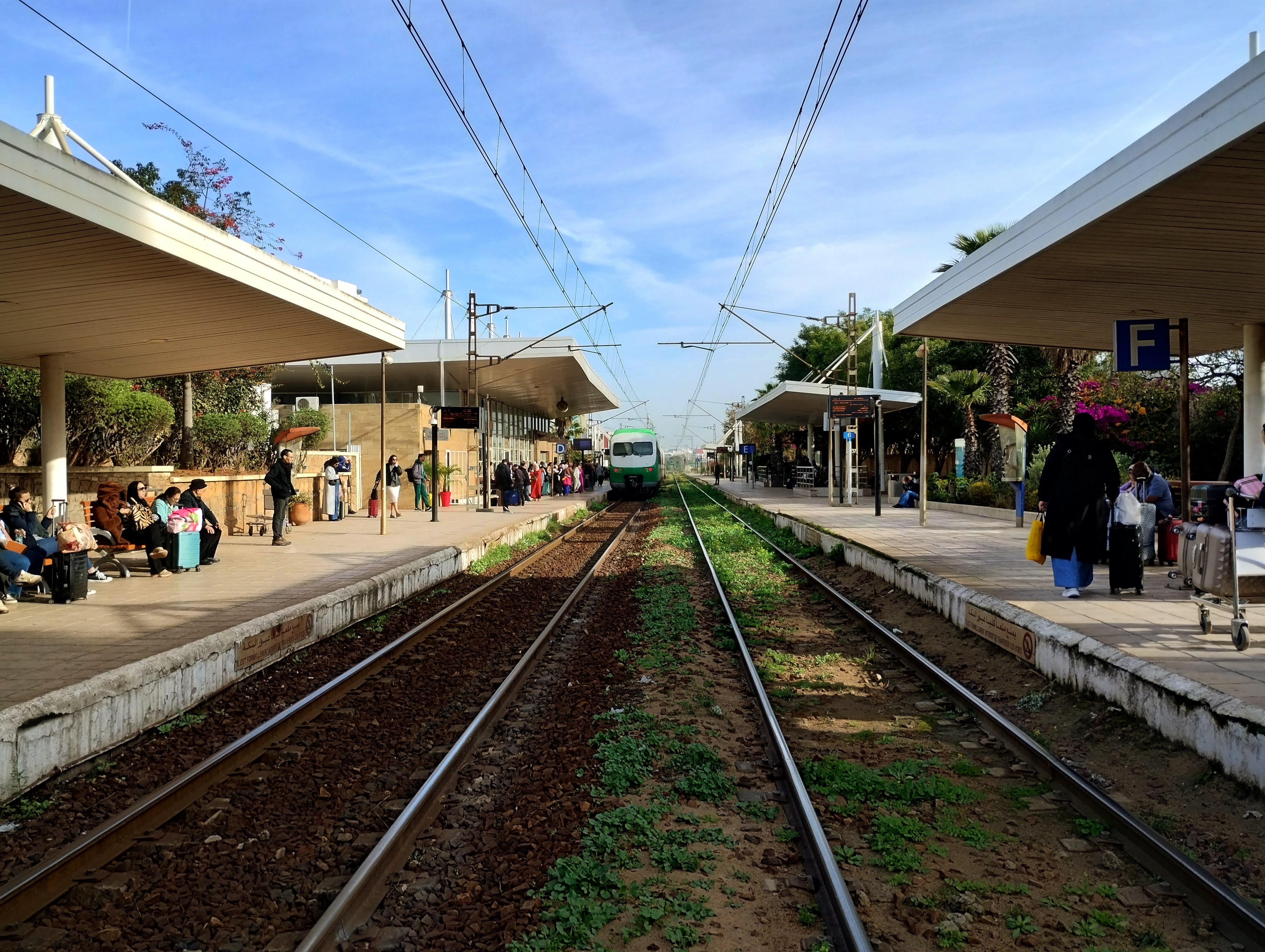 A train station platform on a sunny day, with a green and white train approaching on the tracks. Passengers are visible on both sides of the tracks, waiting on platforms covered by a white canopy. The platform to the right has a sign with a white "F" in a blue square. Overhead electrical wires extend across the tracks.