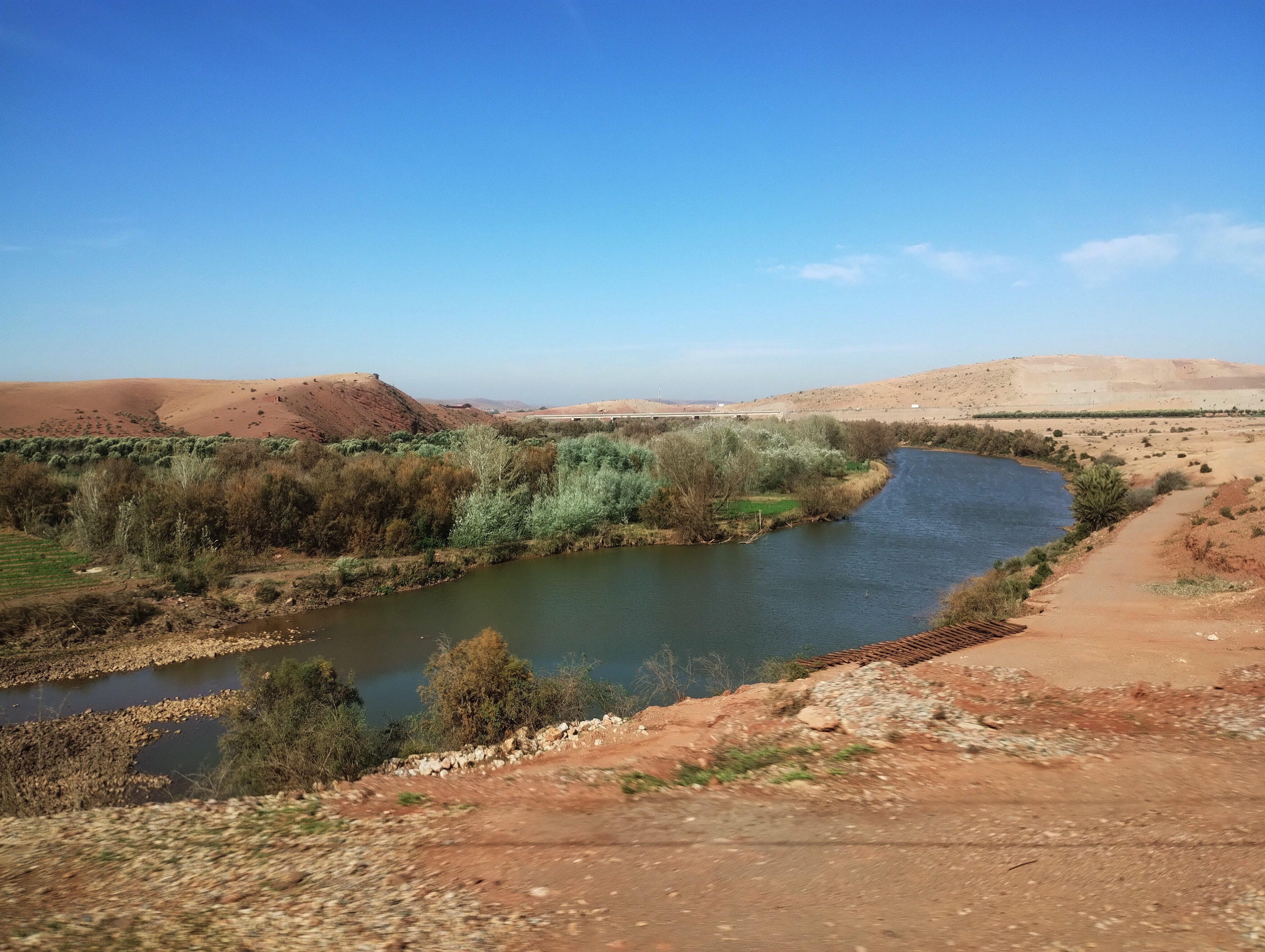 A wide shot of a river flowing through a desert landscape. The river, a dark blue-green, winds its way through the scene, flanked by low-lying vegetation and reddish-brown terrain. The sky is a clear, bright blue. In the background, rolling hills of a similar reddish-brown provide stark contrast to the blue sky. Vegetation, including trees and bushes, lines the riverbanks.
