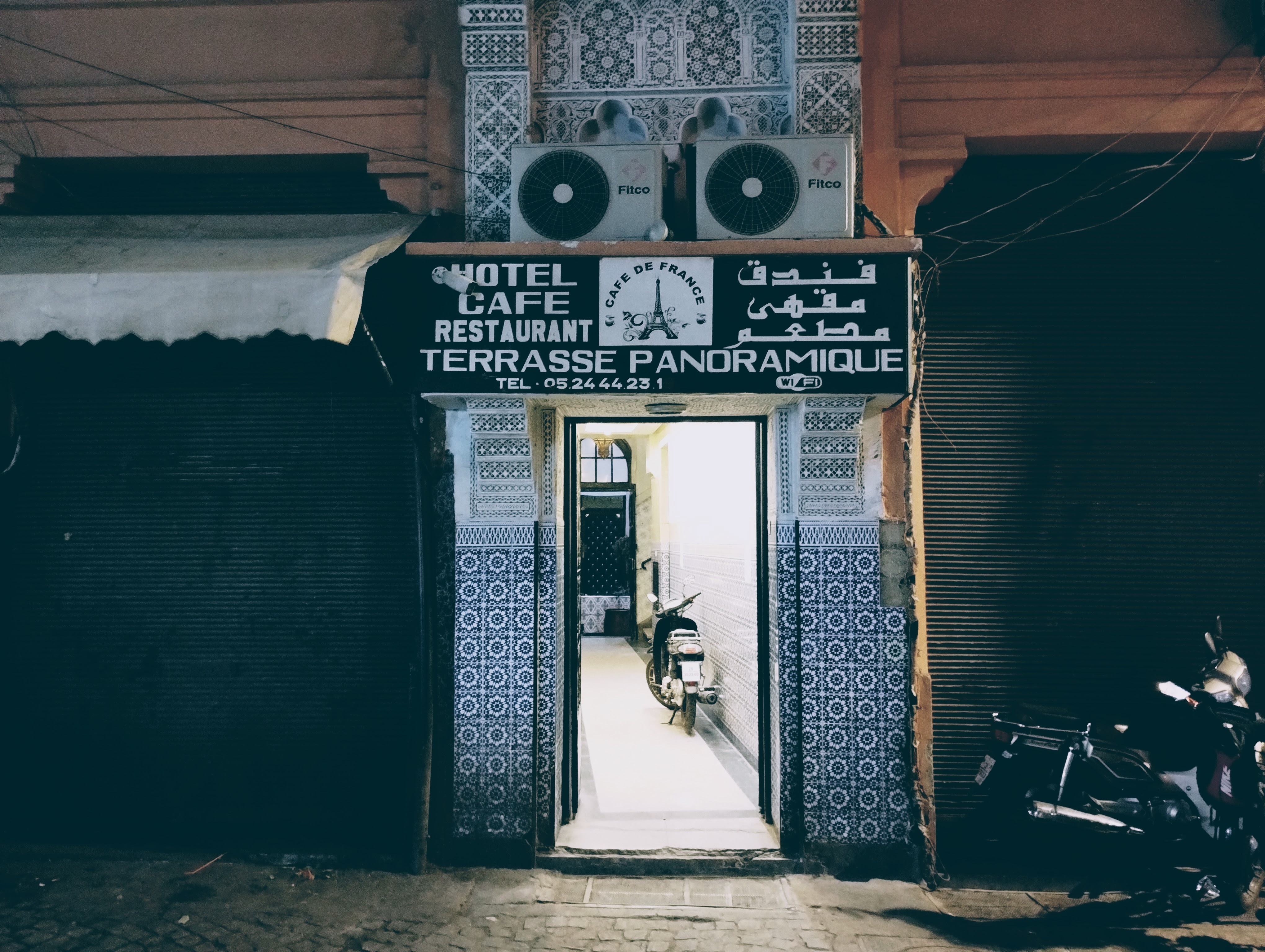 The entrance to the Café de France at night, with signage that reads "HOTEL CAFE RESTAURANT TERRASSE PANORAMIQUE" in Latin and Arabic script. The open door is flanked by zellij tiles. A scooter is parked inside the illuminated doorway.