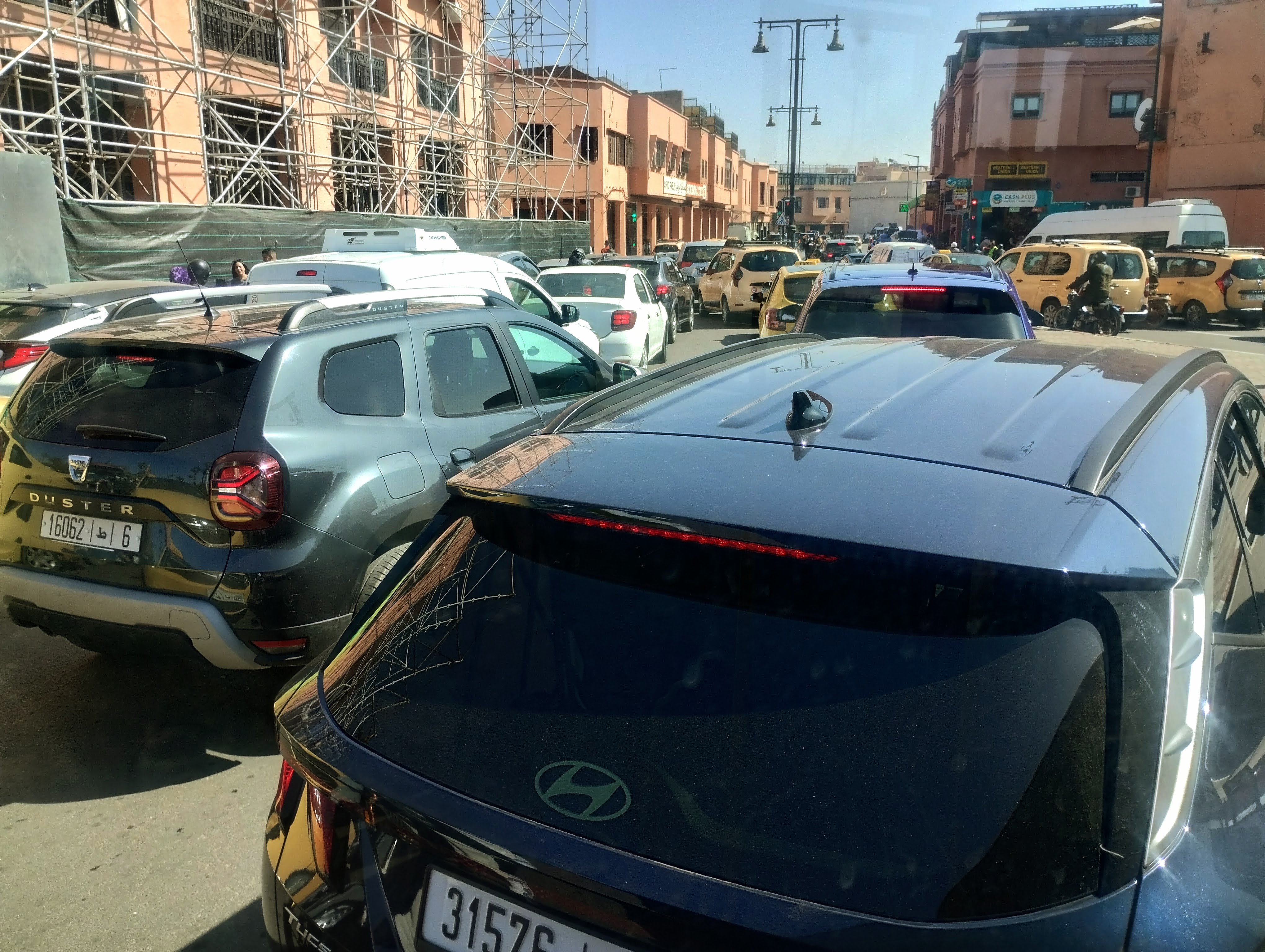 A traffic jam on a sunny day in Marrakech, with various cars and SUVs filling the frame. Further away, rows of cars, including yellow taxis, are packed into the street, and buildings are visible along the road, some under construction. 