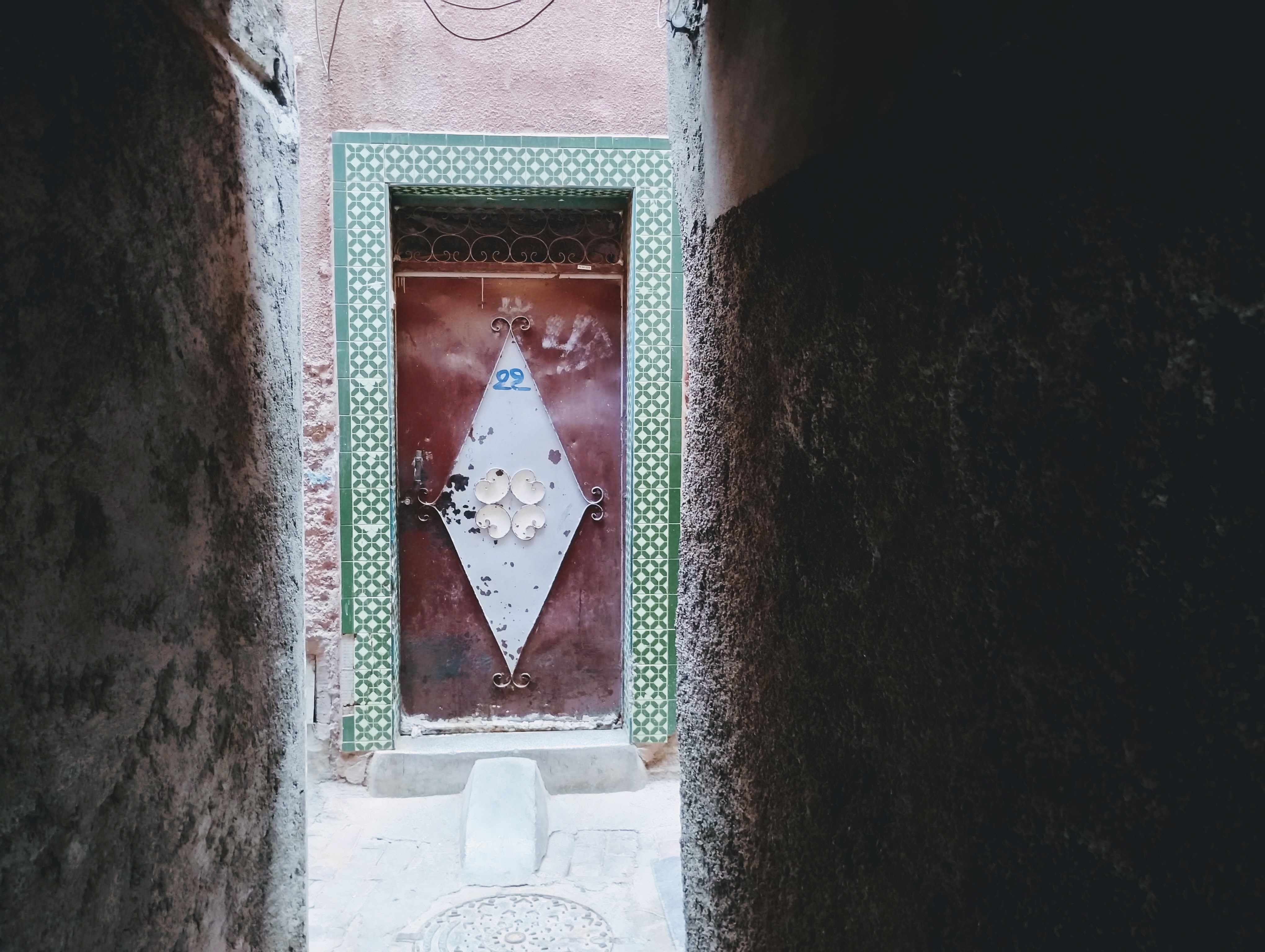 A narrow alleyway opens to a reddish-brown door, framed by green and white tiled trim, and centred with a white diamond shape featuring ornamental metalwork and a small blue "22." The alley walls, dark and textured, frame the scene, while a small concrete block sits at the base of the door.