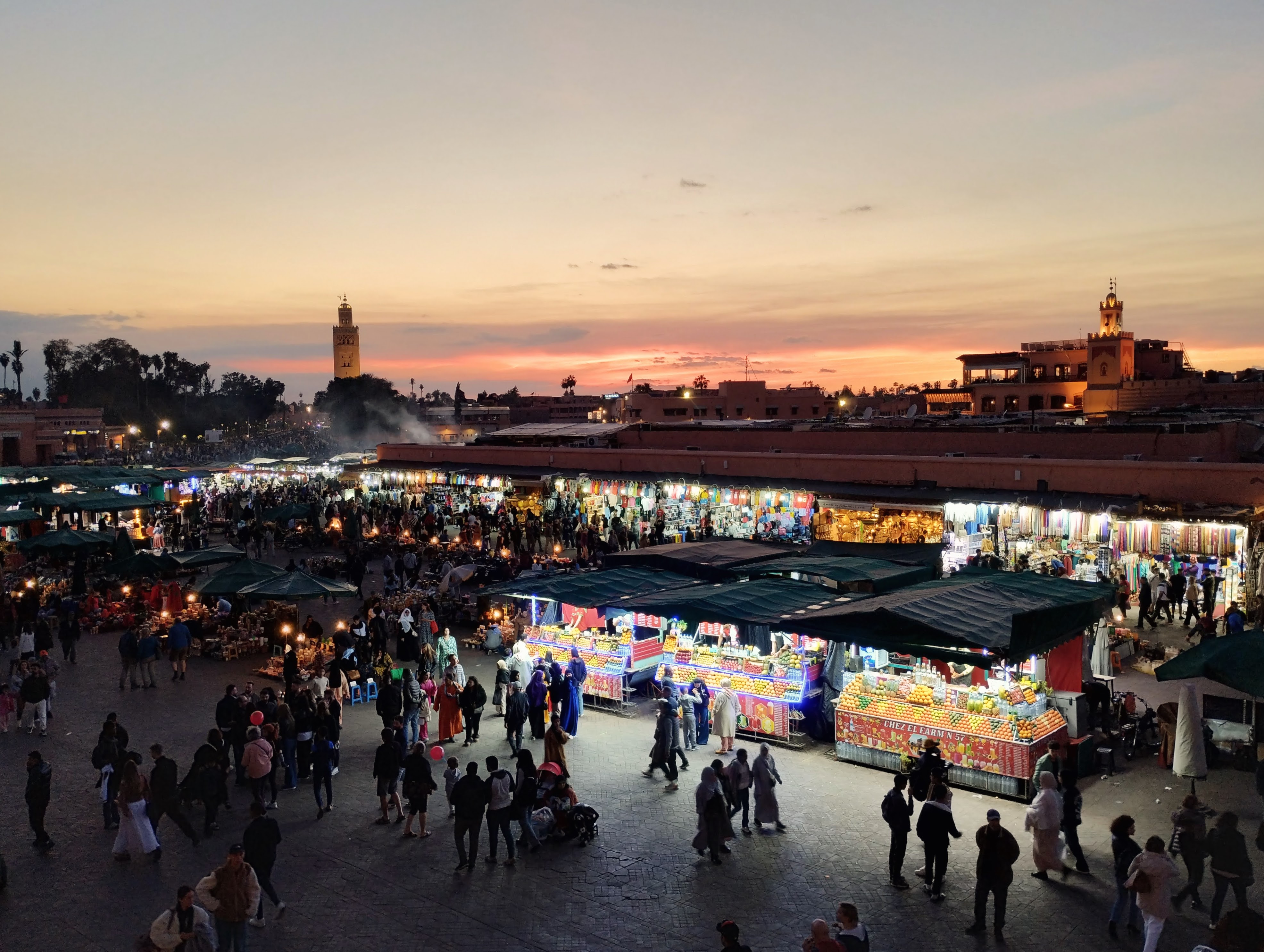 Overhead shot of Jemaa el-Fna square in Marrakesh at dusk, with the Koutoubia Mosque minaret visible in the background and a bustling market scene illuminated by stall lights. Fruit and other goods are displayed at vendors, while crowds of people walk across the open square.