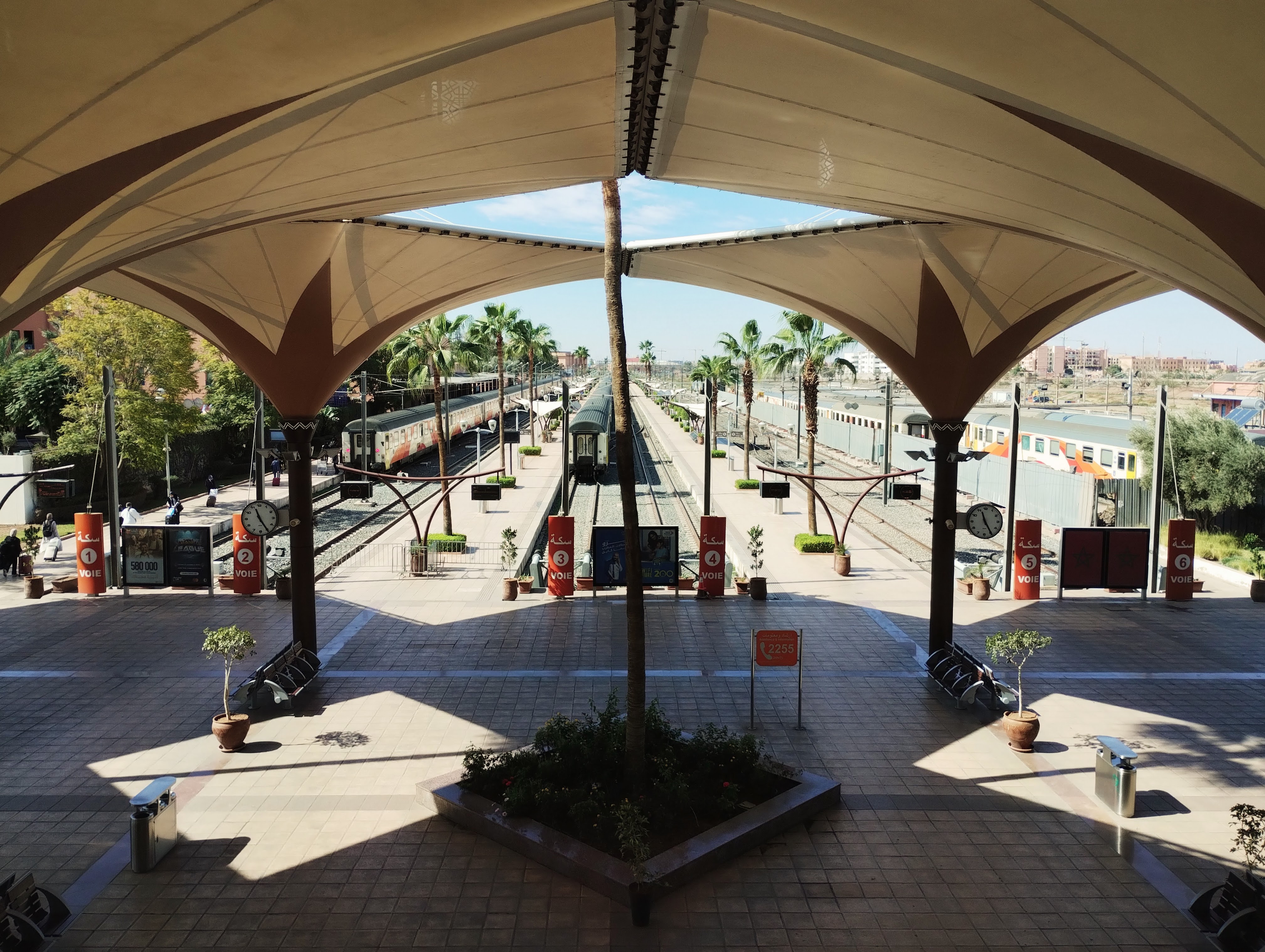A view along train station platforms at Marrakech, framed by a beige and brown canopy. The platforms have multiple tracks with trains on both sides. Palm trees are spaced along the platforms, and the sky is visible through the canopy's opening. There are clocks, benches, and potted plants visible.