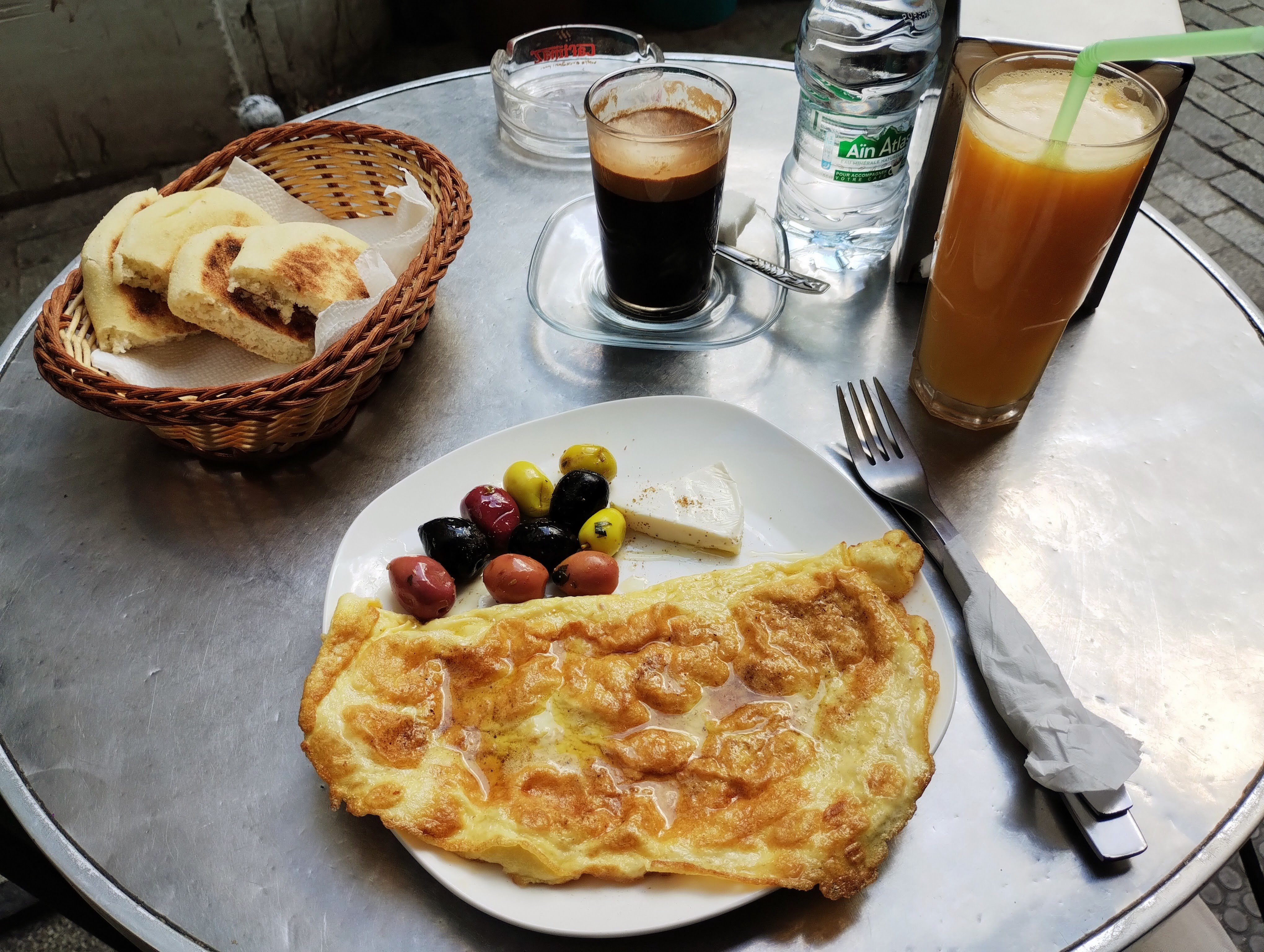 Breakfast of coffee, orange juice, breads, and an omelette with olives on a metal table.