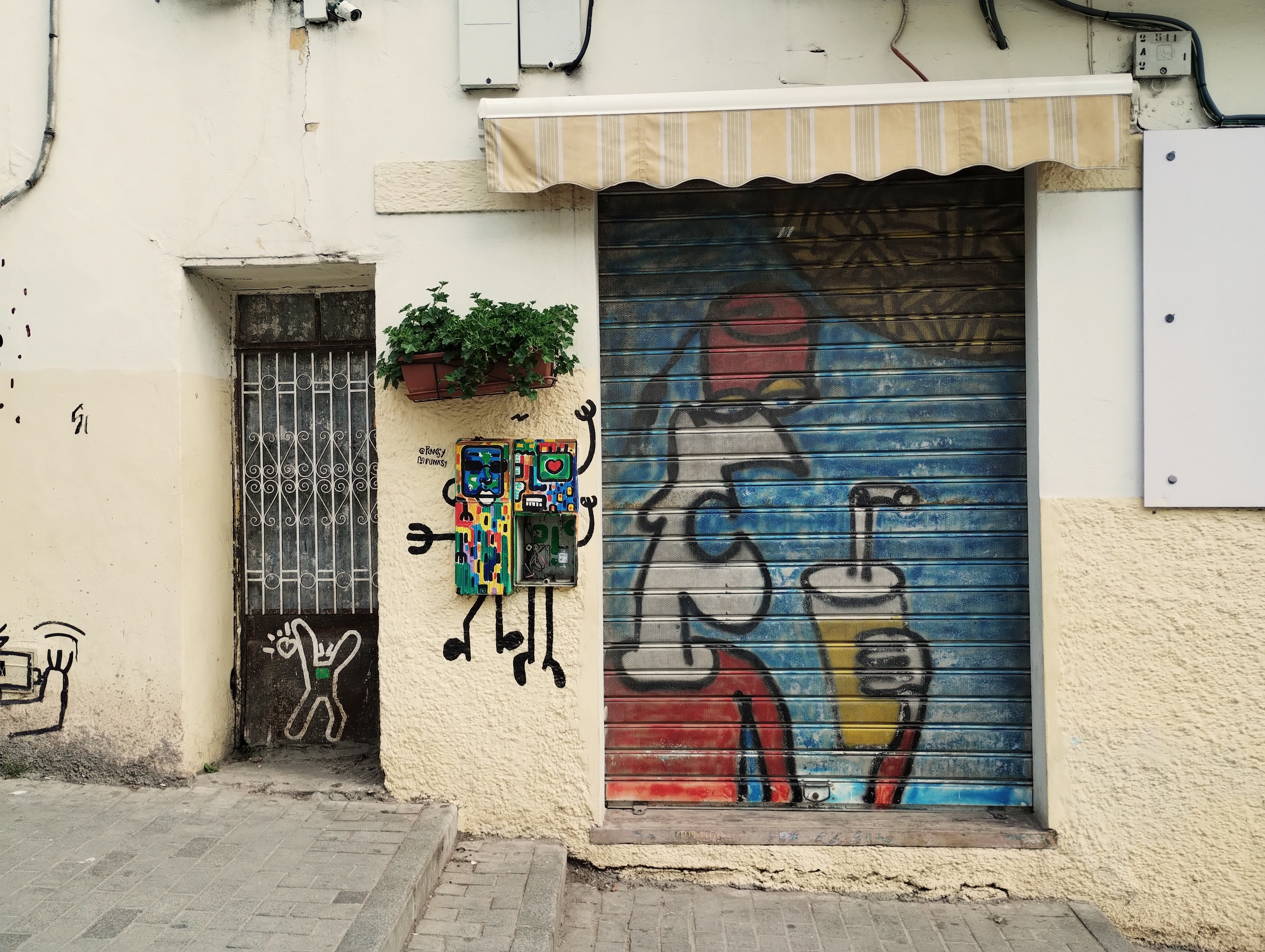 A brown door with white metalwork, two decorated electricity meters, and spray art of a man holding a drink painted onto a metal shutter. 