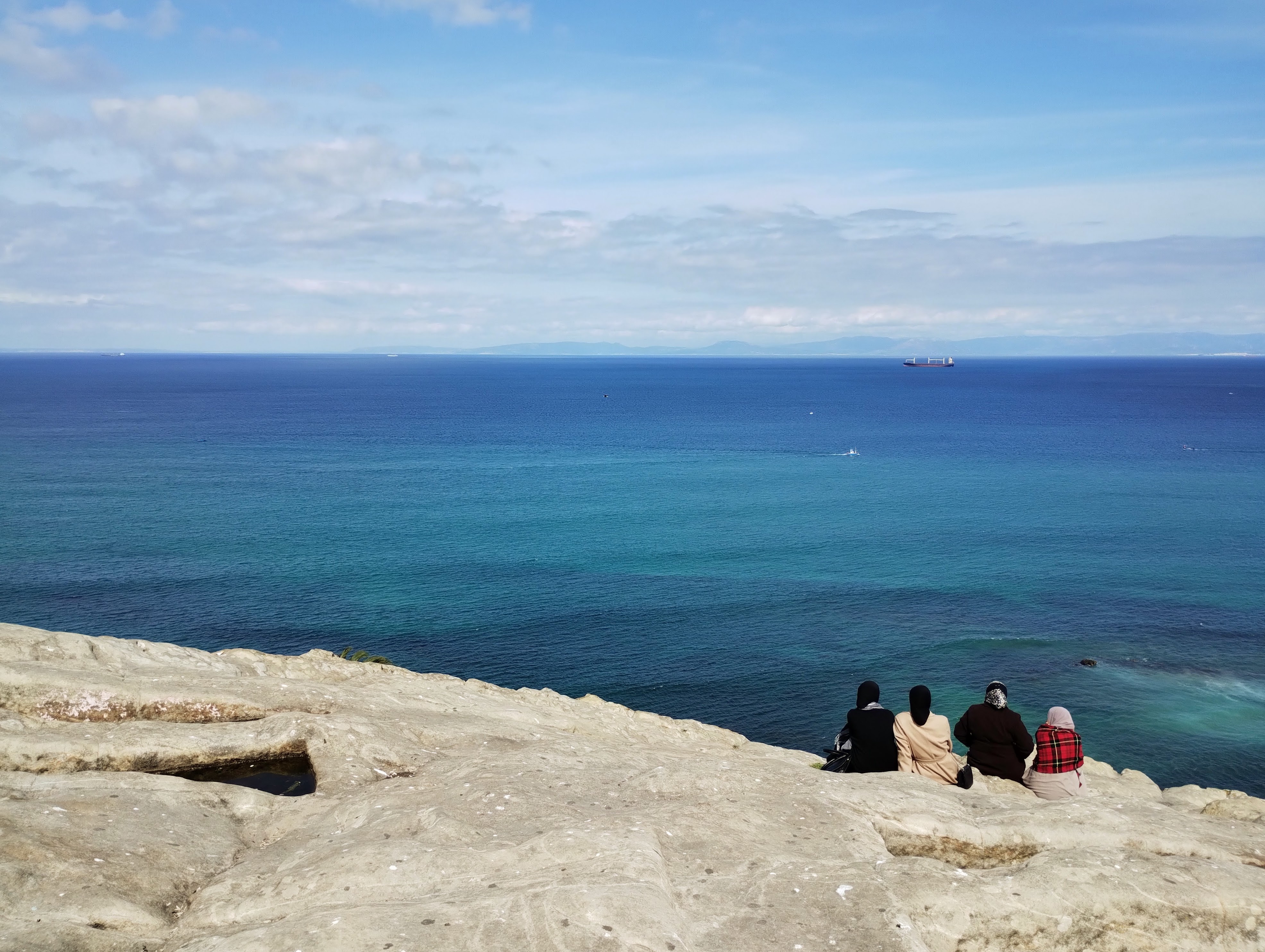 Four women sitting on white rocks look at the outline of Spain across the deep blue trait of Gibraltar.