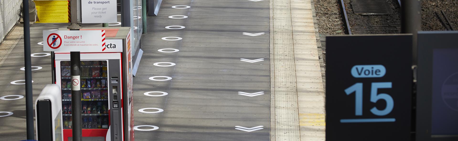 A French train platform shows COVID-19 safety measures with white circles and directional arrows painted on the ground to assist with social distancing. A vending machine with "Danger!" signs and a platform display indicating "Voie 15" is in view.
