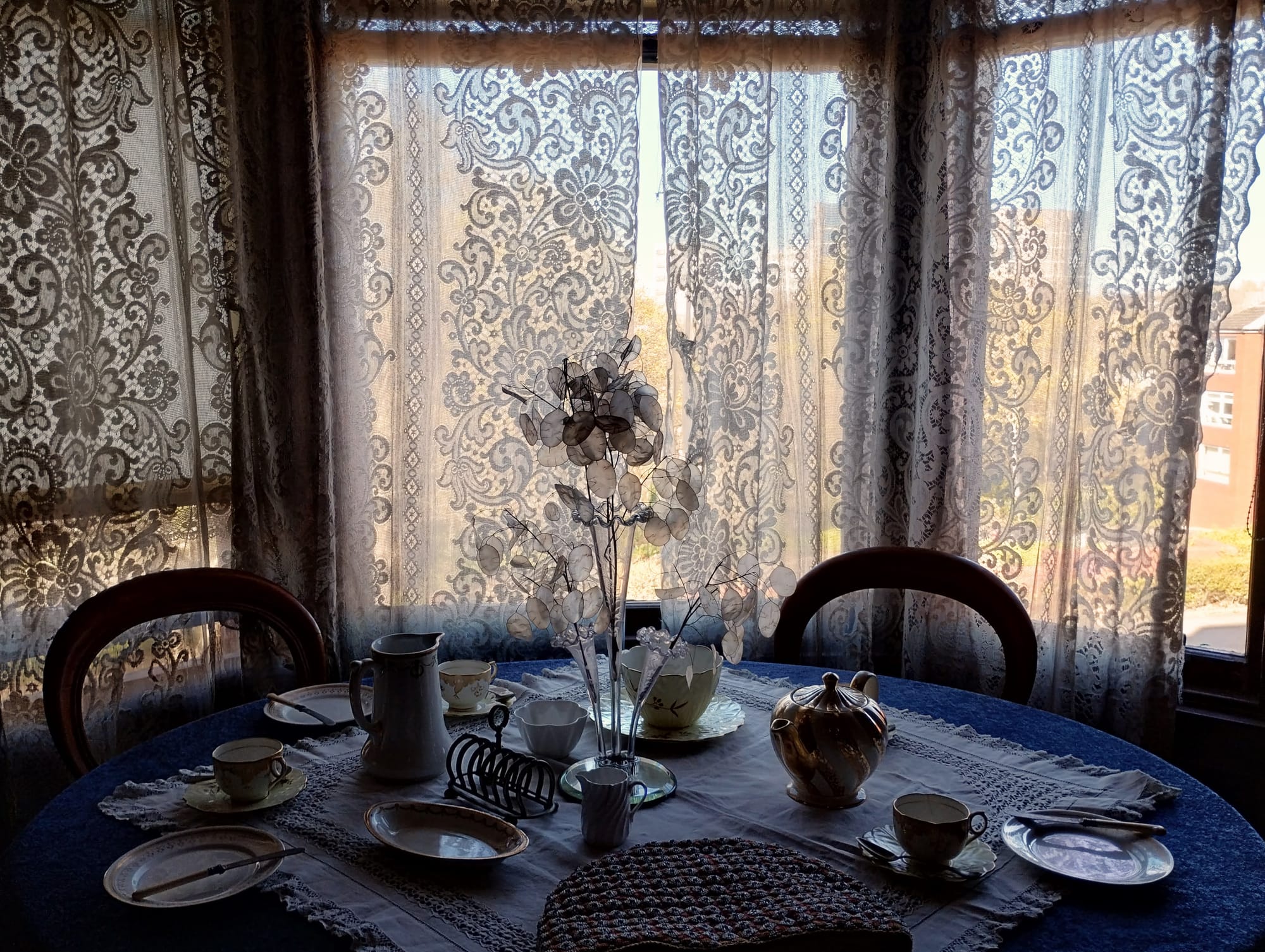 A table set for tea in front of net-curtains in a bay window.