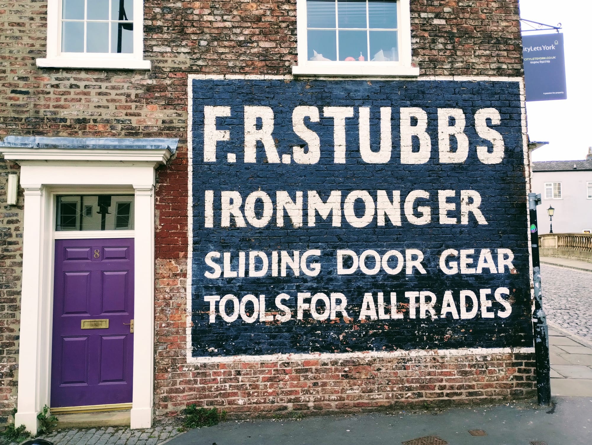 A purple wooden door in a brick wall on which is painted an advert for "F R Stubbs Ironmonger."