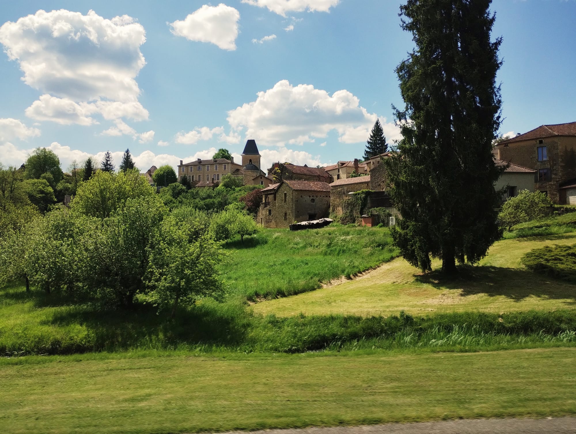 A stone village on a hill in the south-west of France. At the top a church. In the foreground, lush grass and trees.