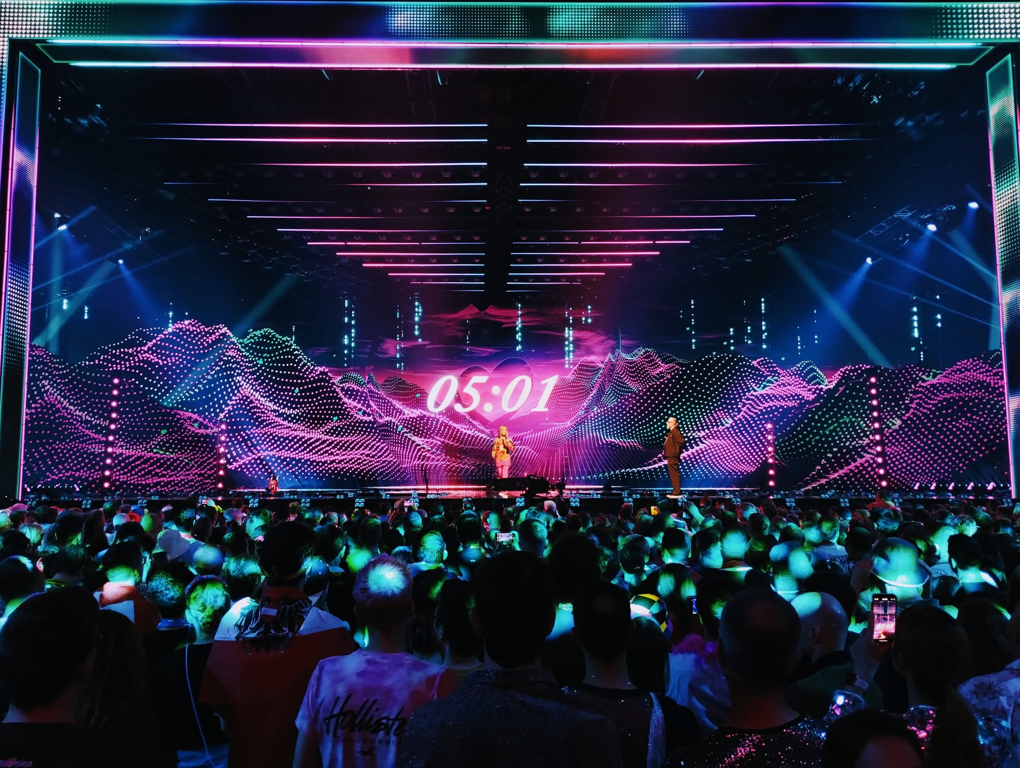Two presenters stand on the stage at the Eurovision Song Contest, beneath a count-down clock.
