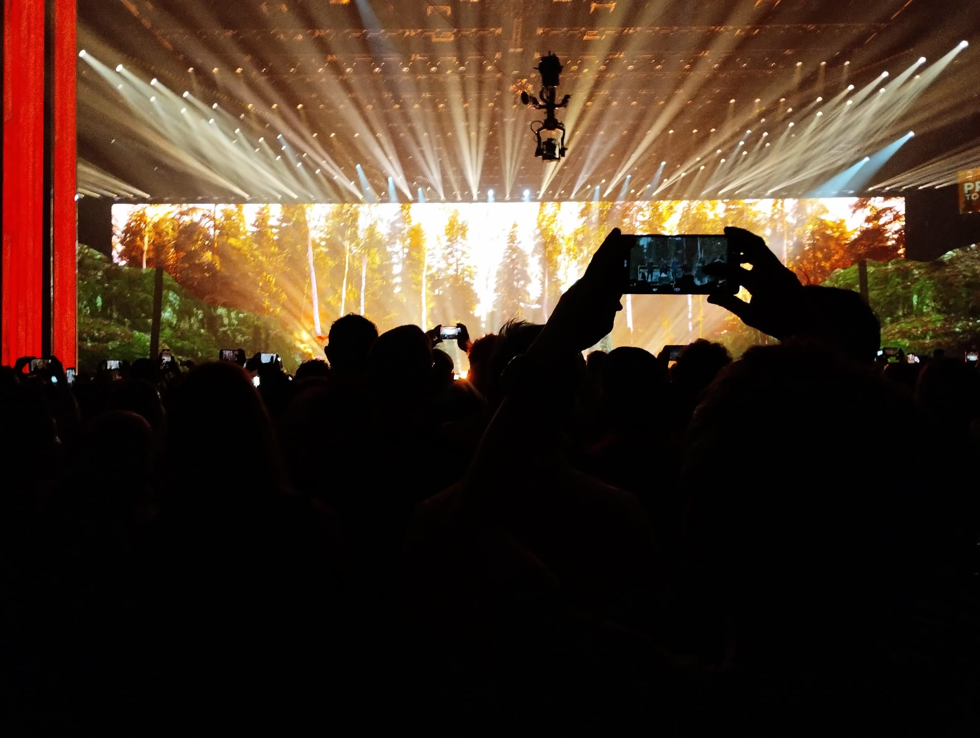 Audience silhouetted against a stage with a large screen displaying a forest scene illuminated by spotlights, with many attendees holding up phones to record the event.