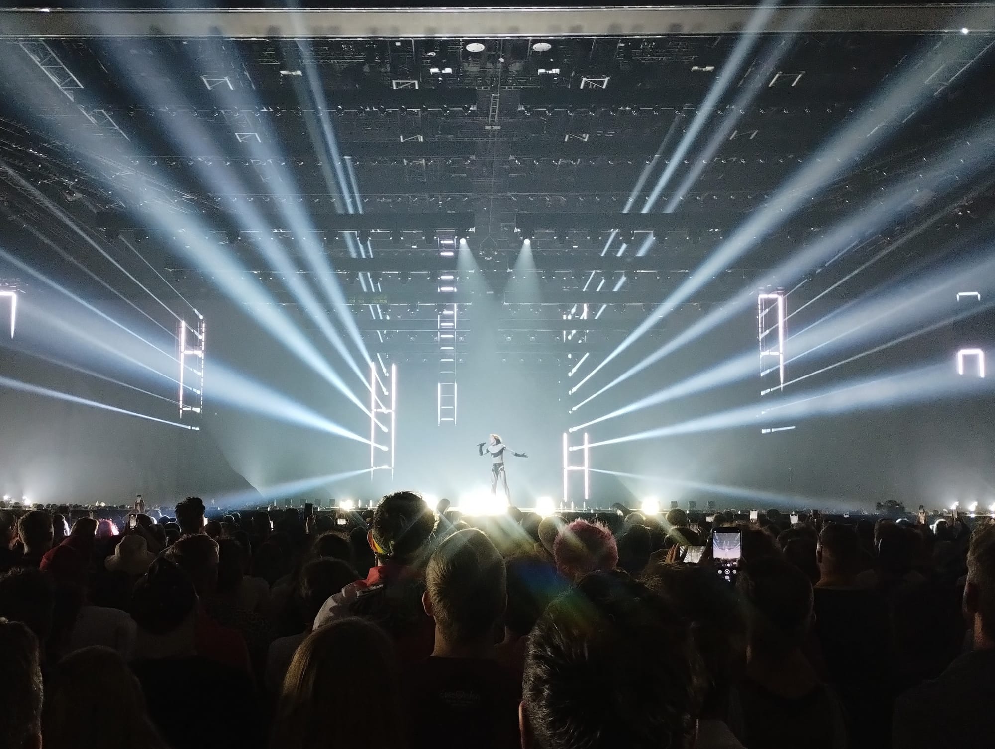 A concert stage bathed in bright white spotlights, with Nemo silhouetted against the light, in front of a large audience with mobile phones raised.