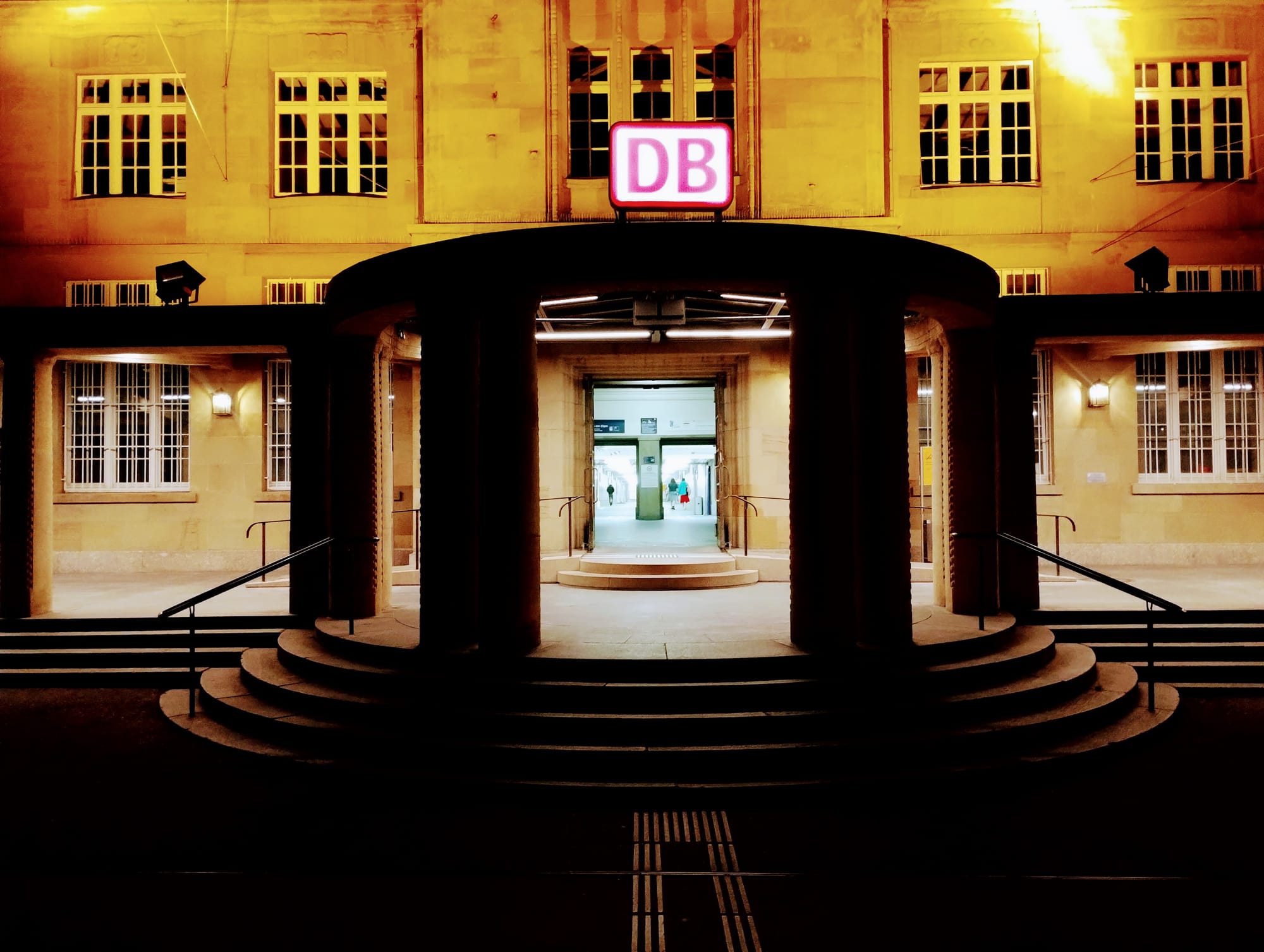 Exterior of a Basel Bad Bahnhof at night, illuminated with warm yellow lighting highlighting architectural details, steps leading up to an arched entrance featuring a pink DB sign, with a visible interior hallway and steps.
