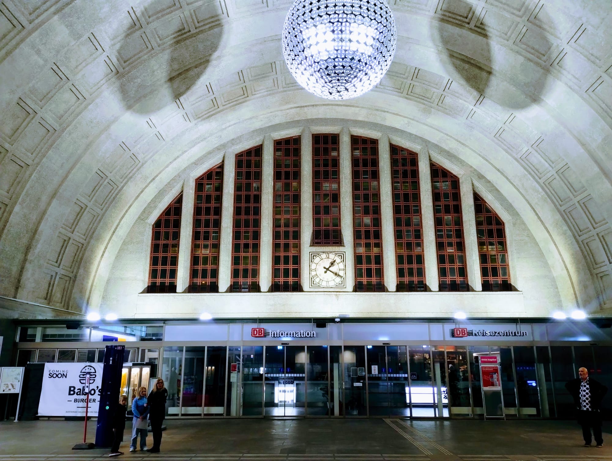 Interior of a train station with a high vaulted ceiling, large arched windows, a decorative chandelier, and a clock. Signage indicates "DB information" and "DB Reisezentrum." People stand near entrances. A "Coming Soon" sign for "Baloo's Burger" is visible.