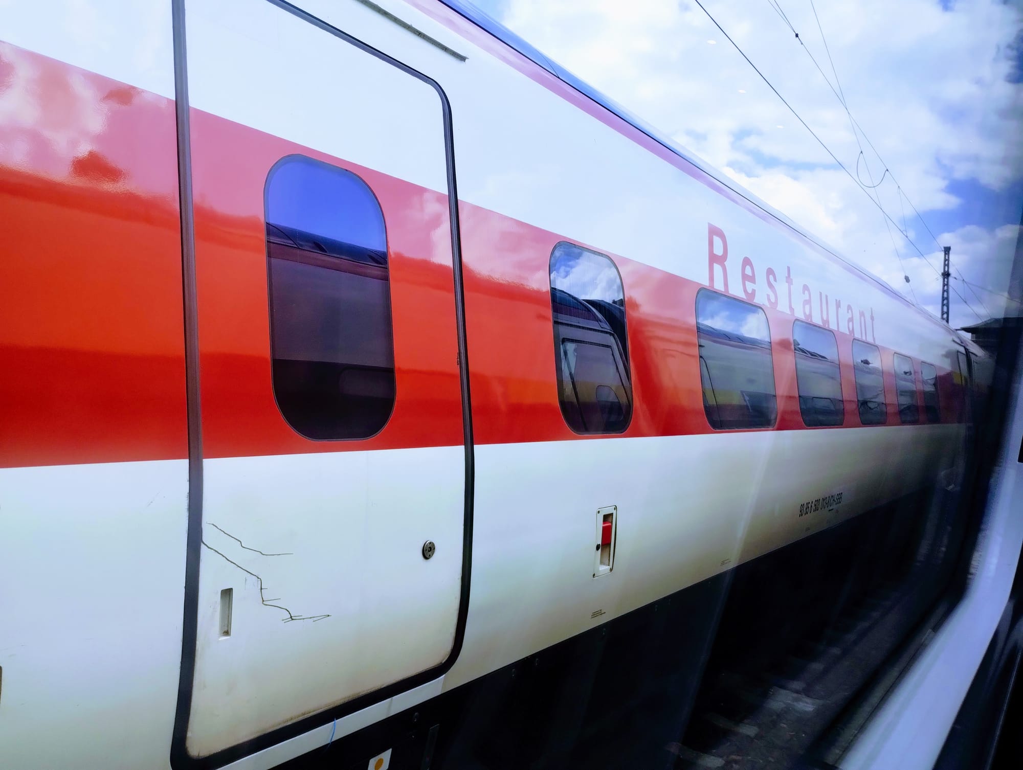 Side view of a red, white, and grey passenger train car, with a "Restaurant" sign visible; the train is viewed through a window on a passing train.