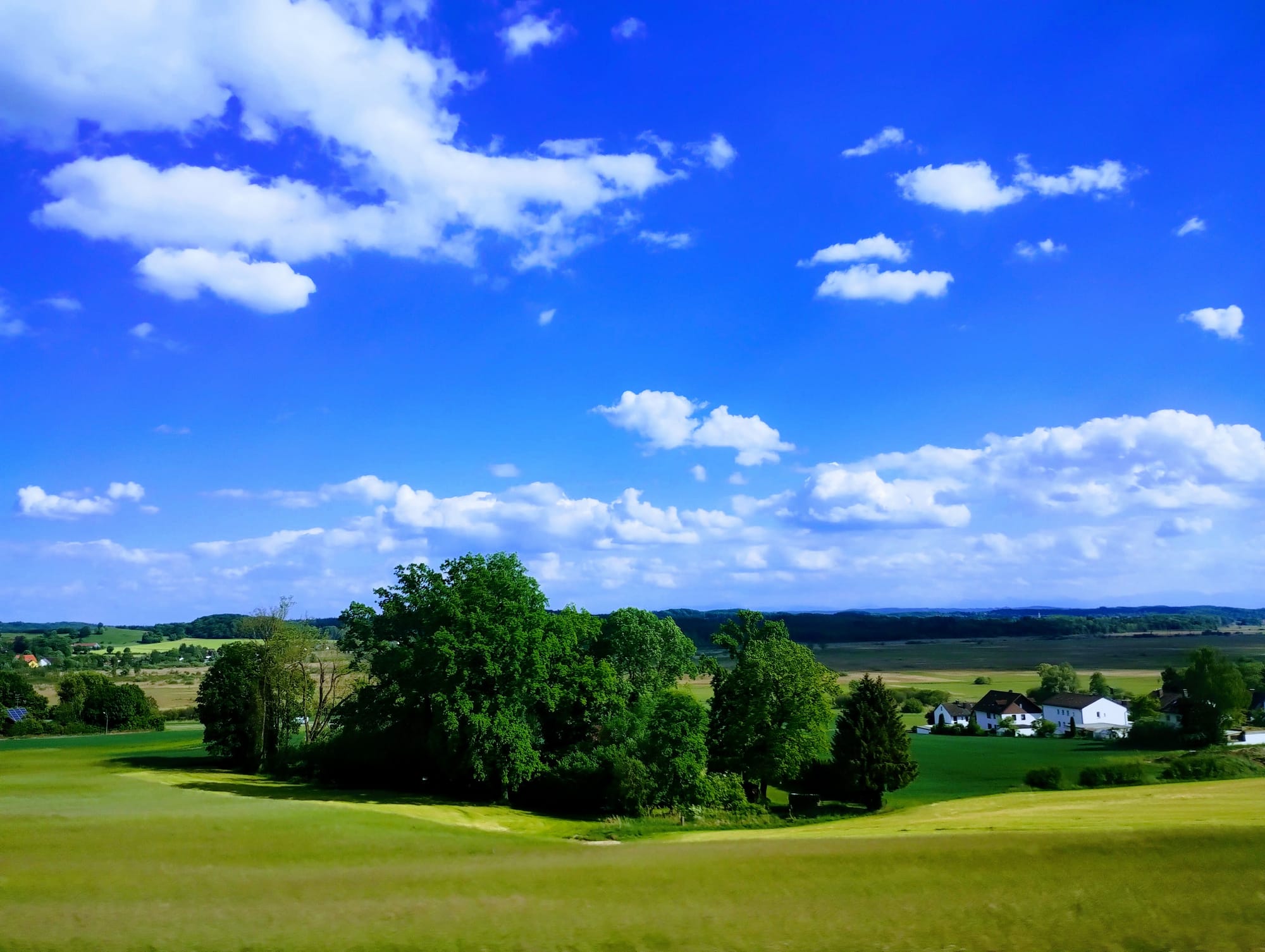 Vast, bright blue sky dotted with cumulus clouds over a rolling green landscape featuring trees, fields, and a cluster of white houses.
