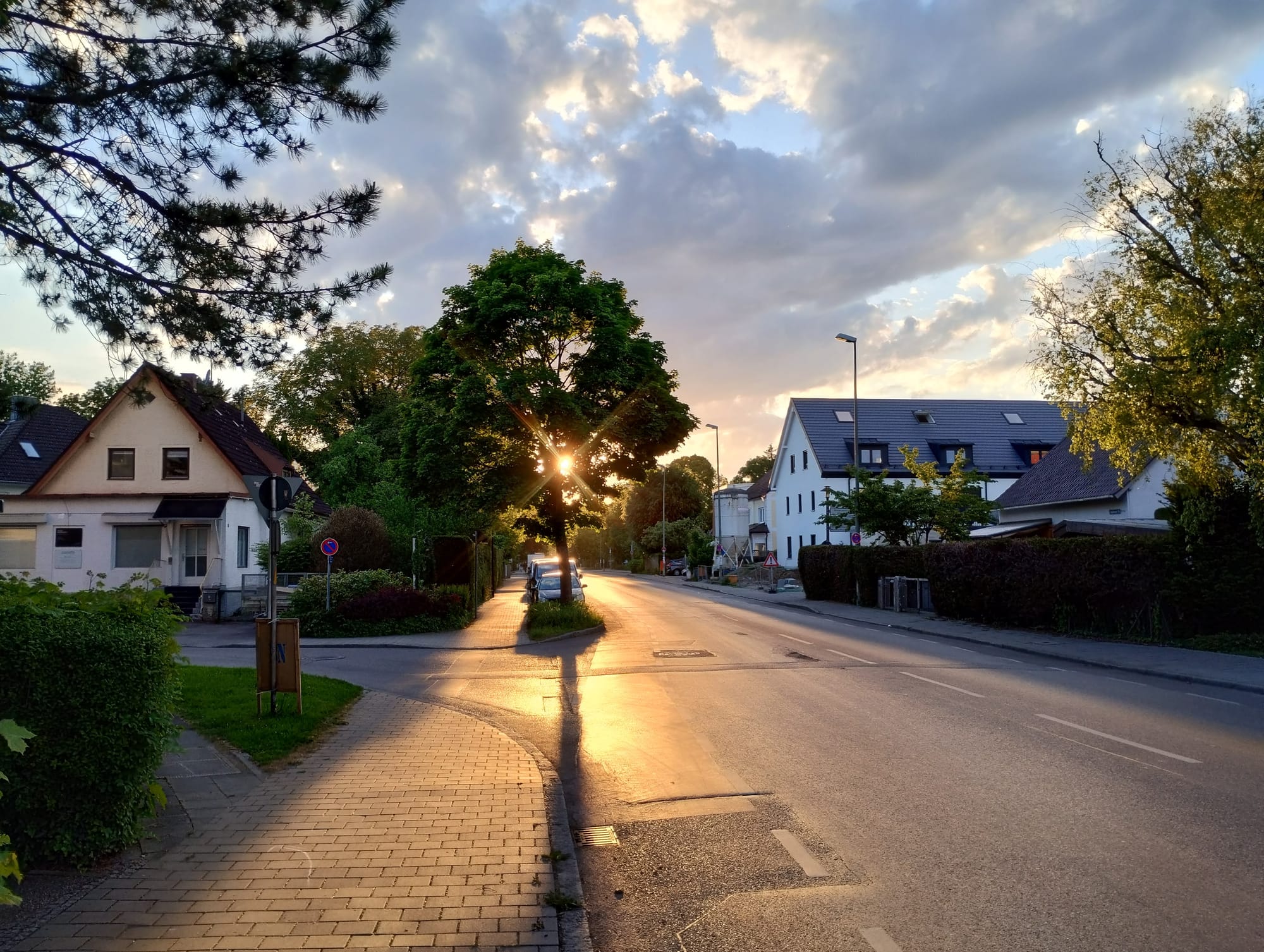 A German street at dusk, bathed in warm sunlight, featuring a road flanked by houses, trees, and a brick sidewalk. Sunlight streams through a large tree in the center, illuminating the asphalt and creating long shadows. The sky is filled with textured clouds, lit by the setting sun.