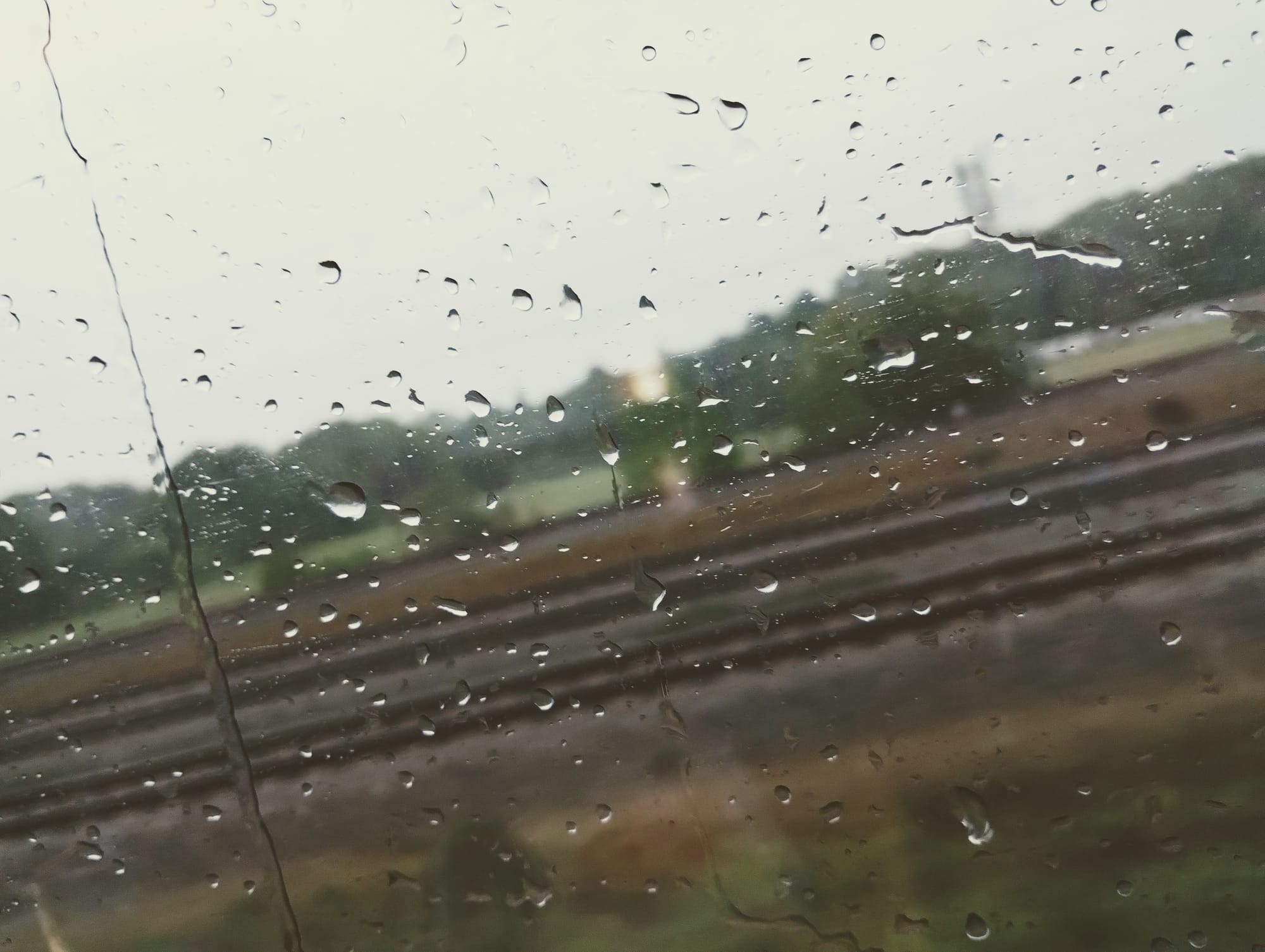 Raindrops on a window, obscuring a blurred view of train tracks, green foliage, and a distant building against a muted grey sky.