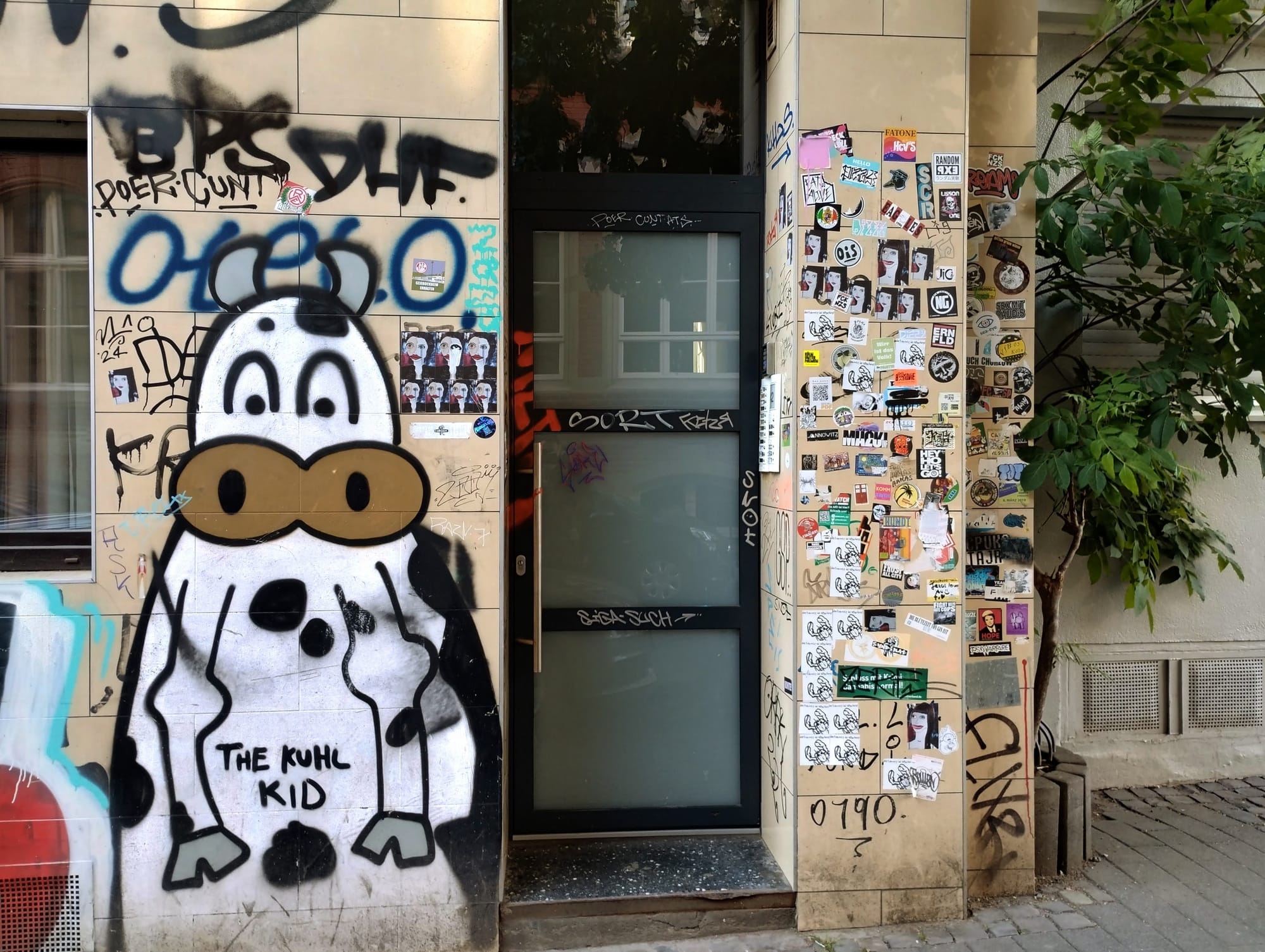 A vibrant street scene featuring a building facade with a large cow-like cartoon mural reading "THE KUHL KID", surrounded by graffiti and stickers, next to a frosted glass door and a small tree.