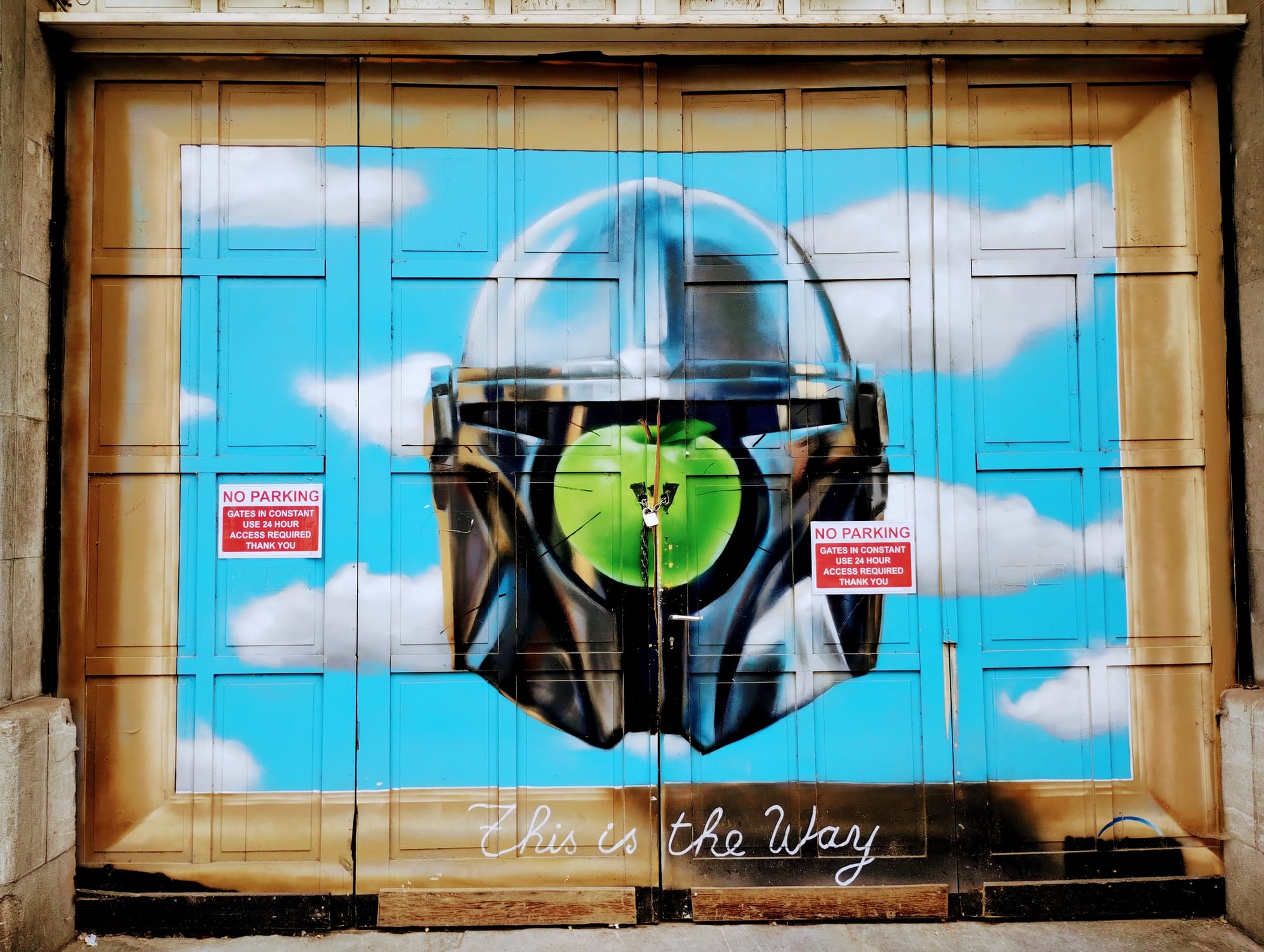 Street art mural on a garage door depicting a chrome Mandalorian helmet with a Magritte apple as the visor against a sky blue backdrop with fluffy white clouds; framed by a golden-brown border; "This is the Way" script below; two "No Parking" signs on either side.