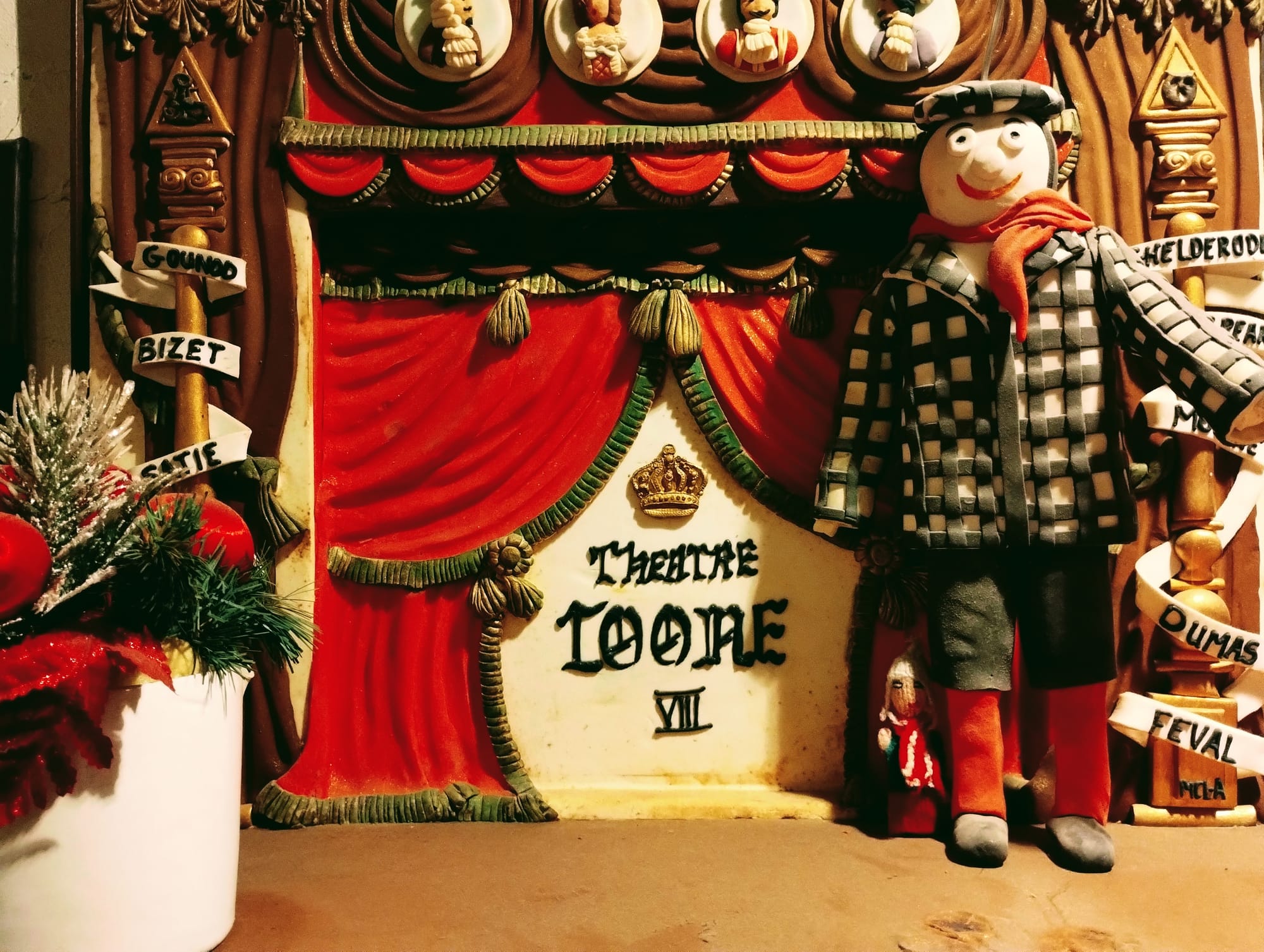 Finger puppet stands in the shadow of a bigger puppet in a check coat on a mini version of the Theatre Toone VIII stage. The stage features red curtains and decorative elements like a crown and small figures of people on top. To the left is a white vase with Christmas decorations, and to the right are pillars with names on ribbons.