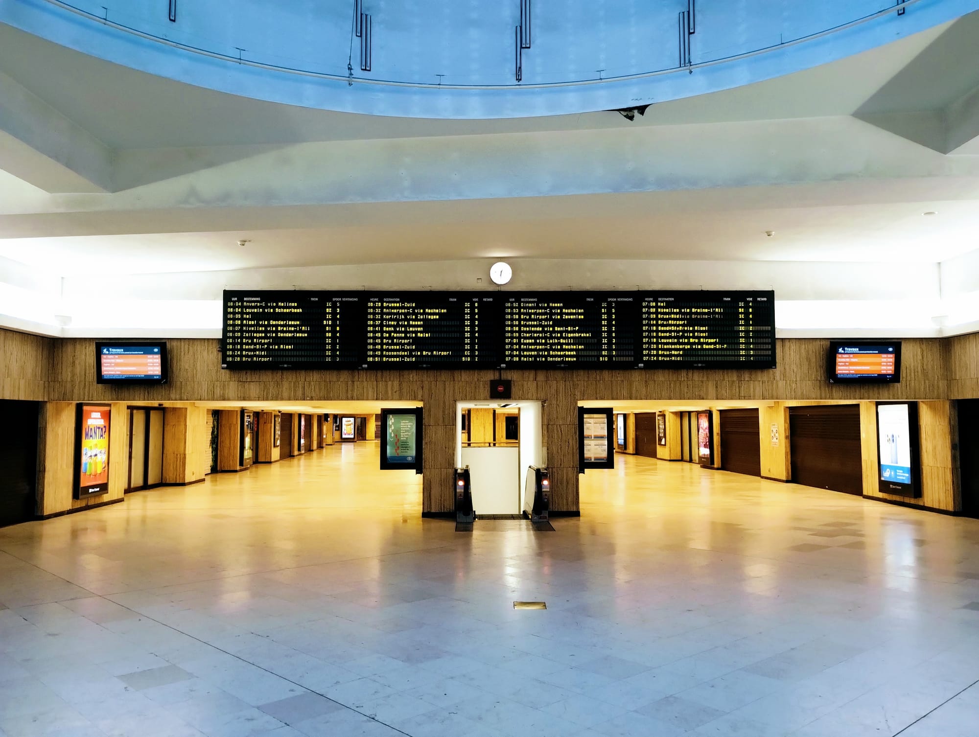 An empty train station concourse featuring large departure boards, illuminated hallways, and a smooth, reflective floor, viewed from a central perspective.