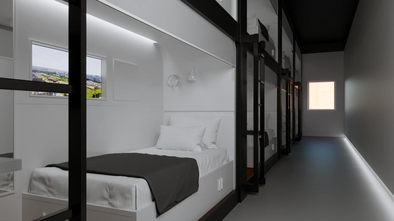 Interior view of a minimalist capsule hostel, bunk beds in white capsules lining the walls, each with a bed, pillow, blanket, and individual window with a photograph of a city view.