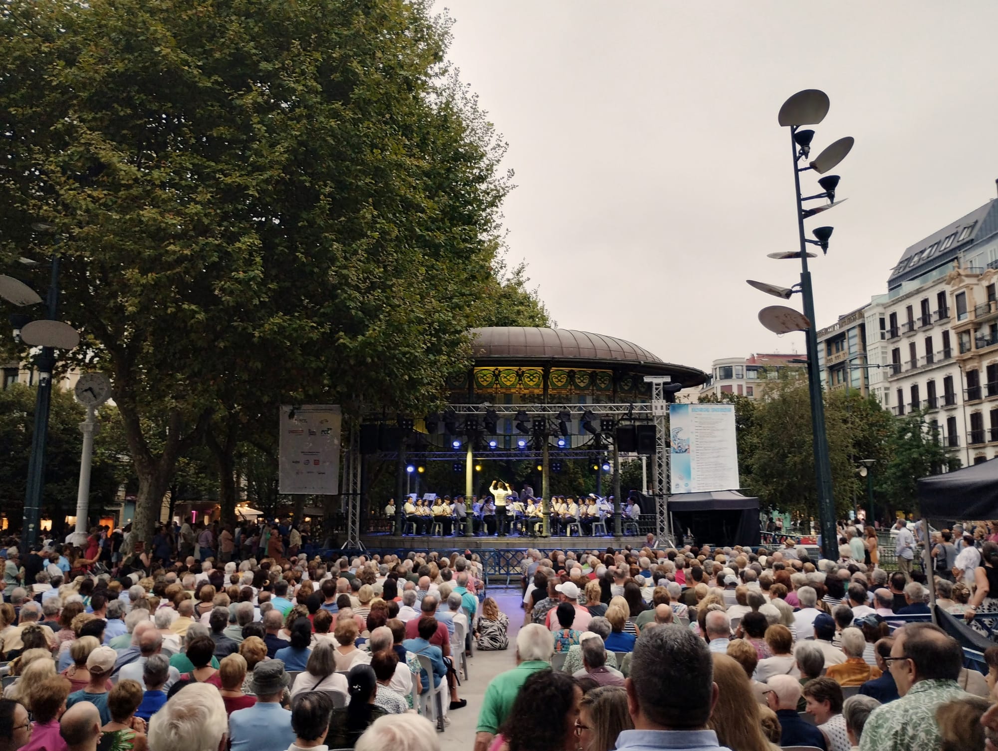 A large audience sits in front of a bandstand listening to a brass band concert. The conductor has his harms in the air.