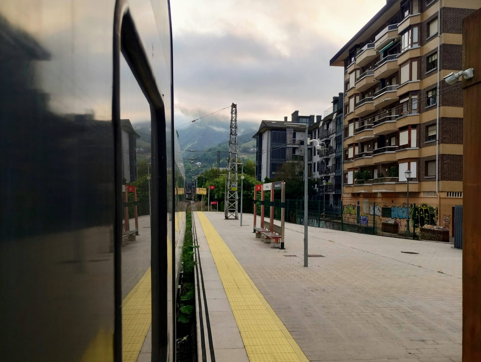 View from a train; on the left, the dark, reflective side of the train. On the right side of the frame, the platform and buildings, including a tall building with balconies and a brick facade. In the distance, mountains are visible through the low cloud.