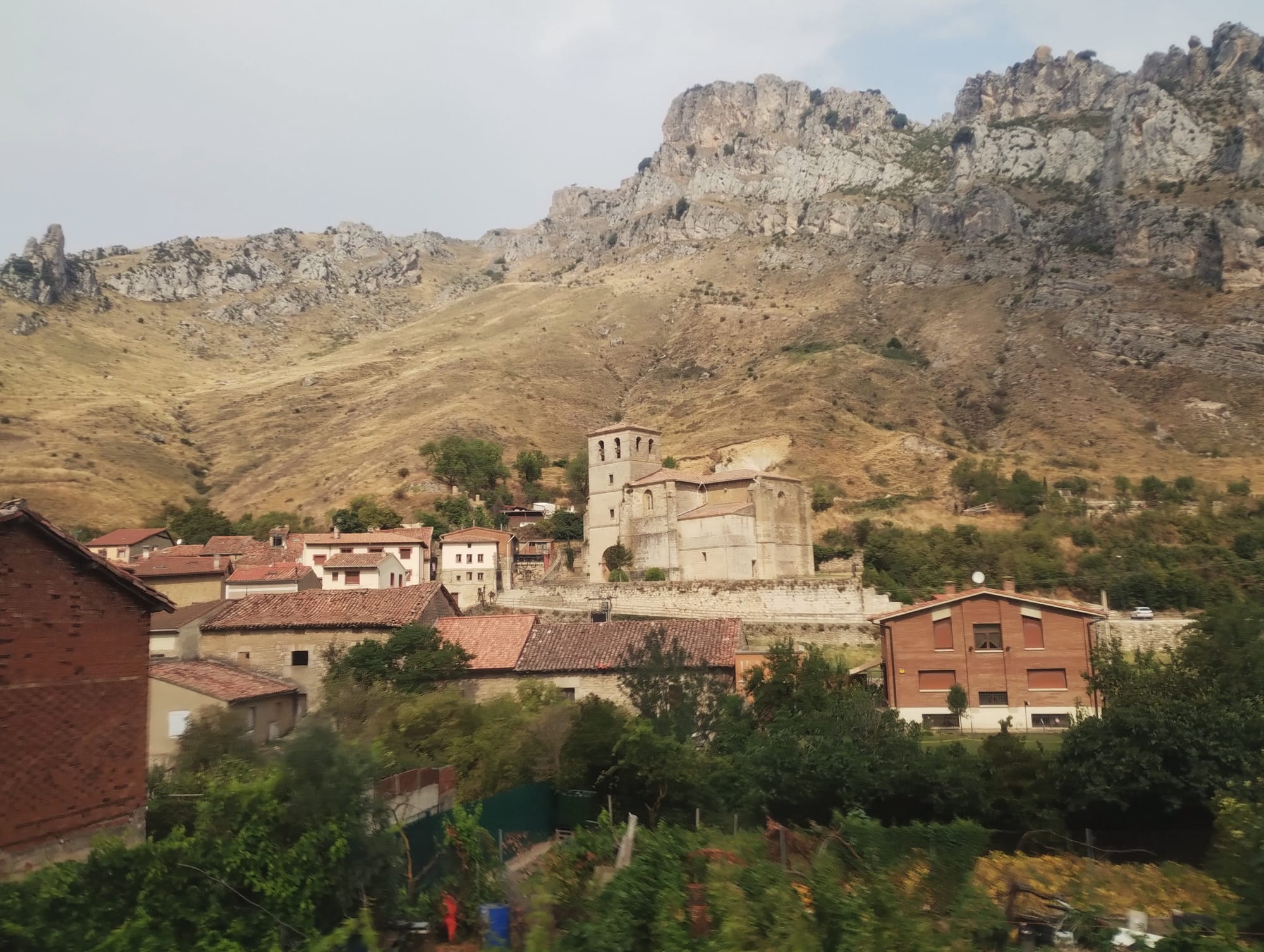 A village nestled at the base of a large, craggy mountain. The village features numerous buildings with reddish-brown tiled roofs and a prominent stone church with a bell tower. The mountain behind dominates the village, which is dotted with green trees and vegetation.