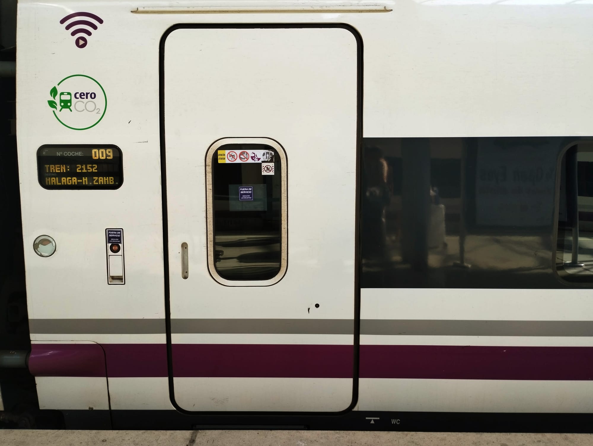 Close-up of a white train car door with a small window, displaying a digital panel showing "009" and the train details, along with informational stickers and a Wi-Fi symbol.